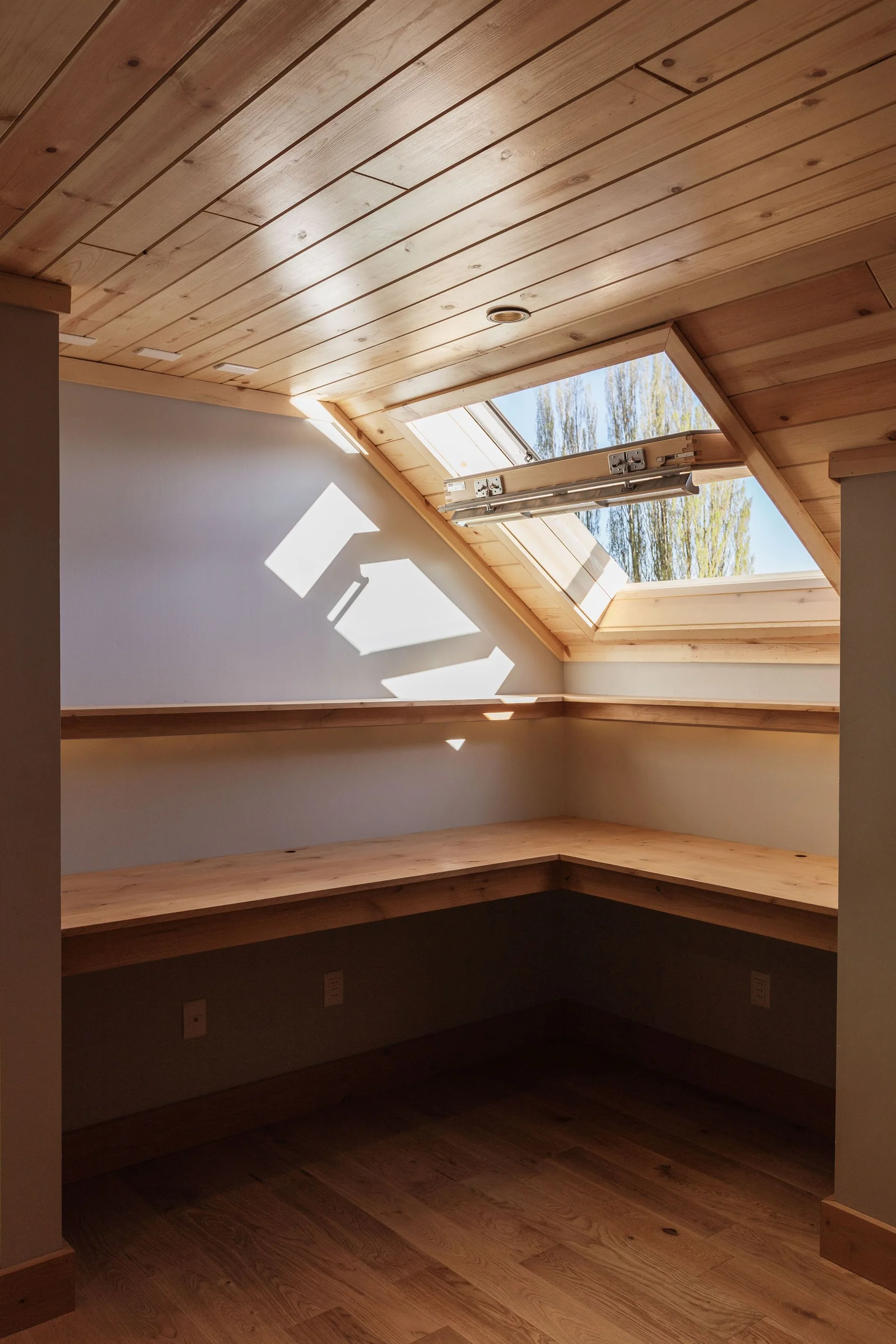 An empty L-shaped wooden desk and floating shelf built into an attic corner with a sloped wood-plank ceiling and a bright skylight.
