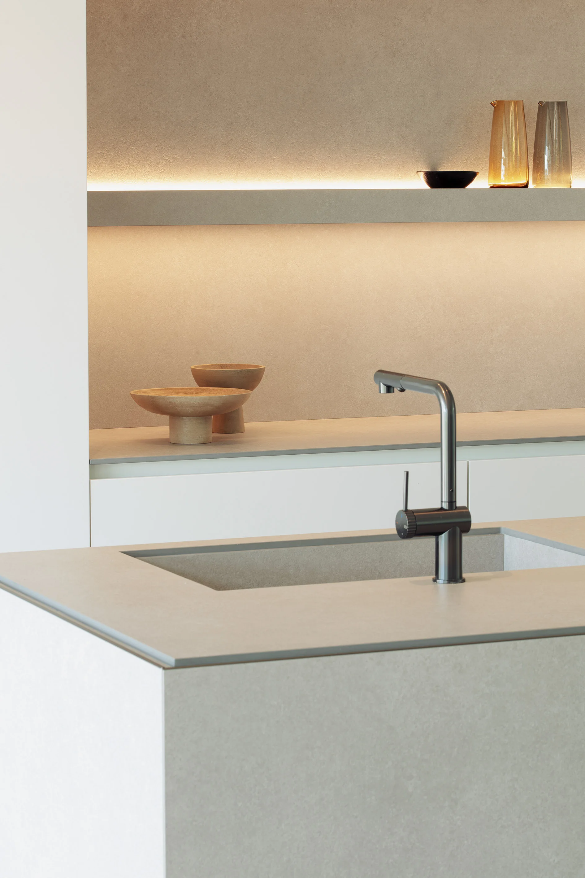 A minimalist kitchen features a stone-look island with an integrated sink, white base cabinets, and a backlit beige shelf displaying glassware.