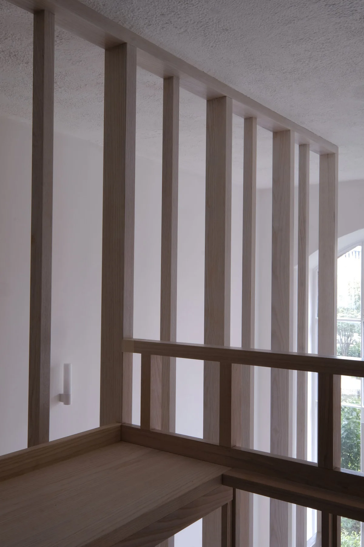 A minimalist partition wall made of vertical light wood slats stands on a staircase landing next to a white wall and a window.