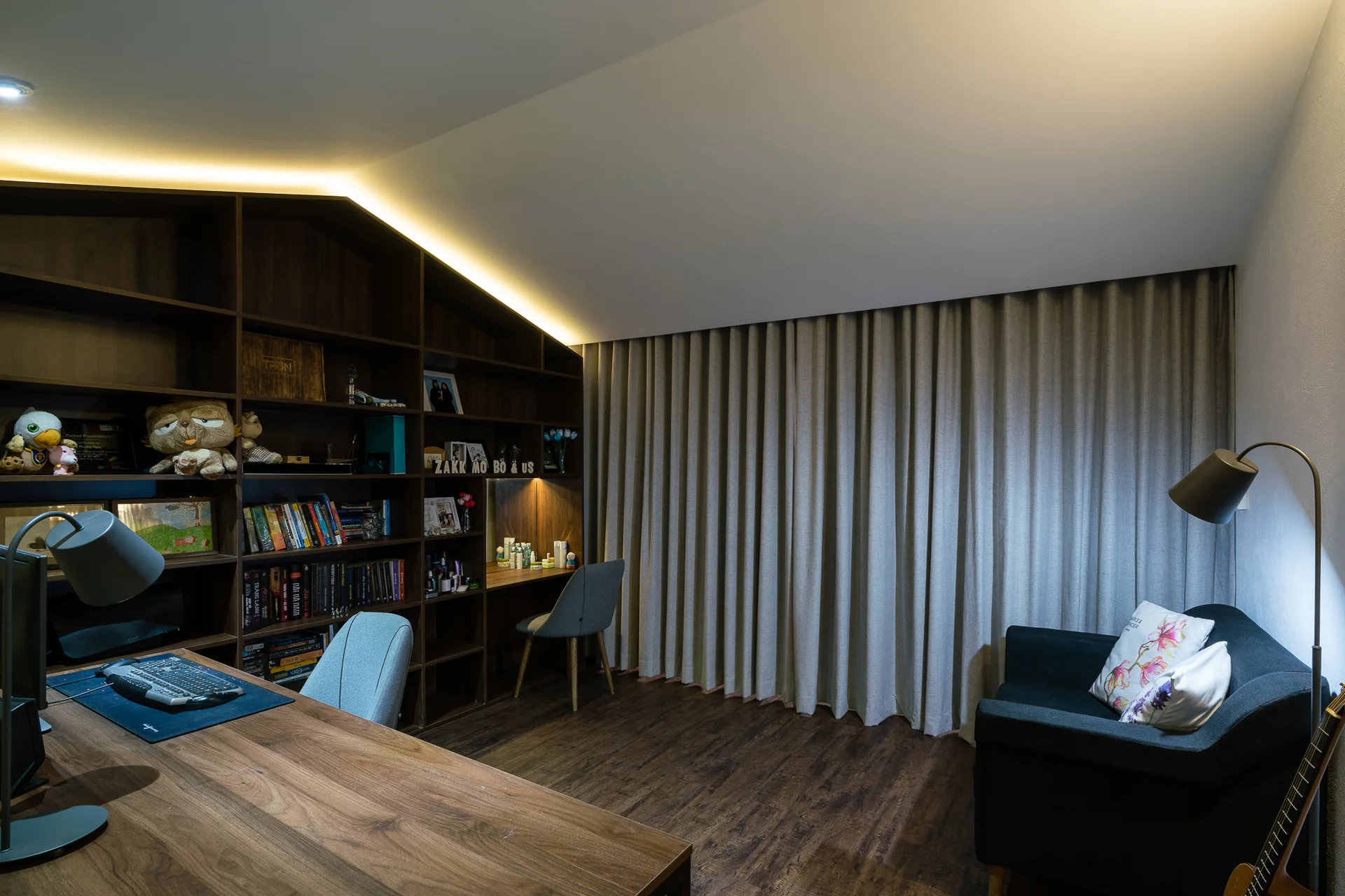 A modern home office featuring a sloped ceiling, dark wood built-in bookshelves with integrated lighting, a wooden desk, and grey curtains.