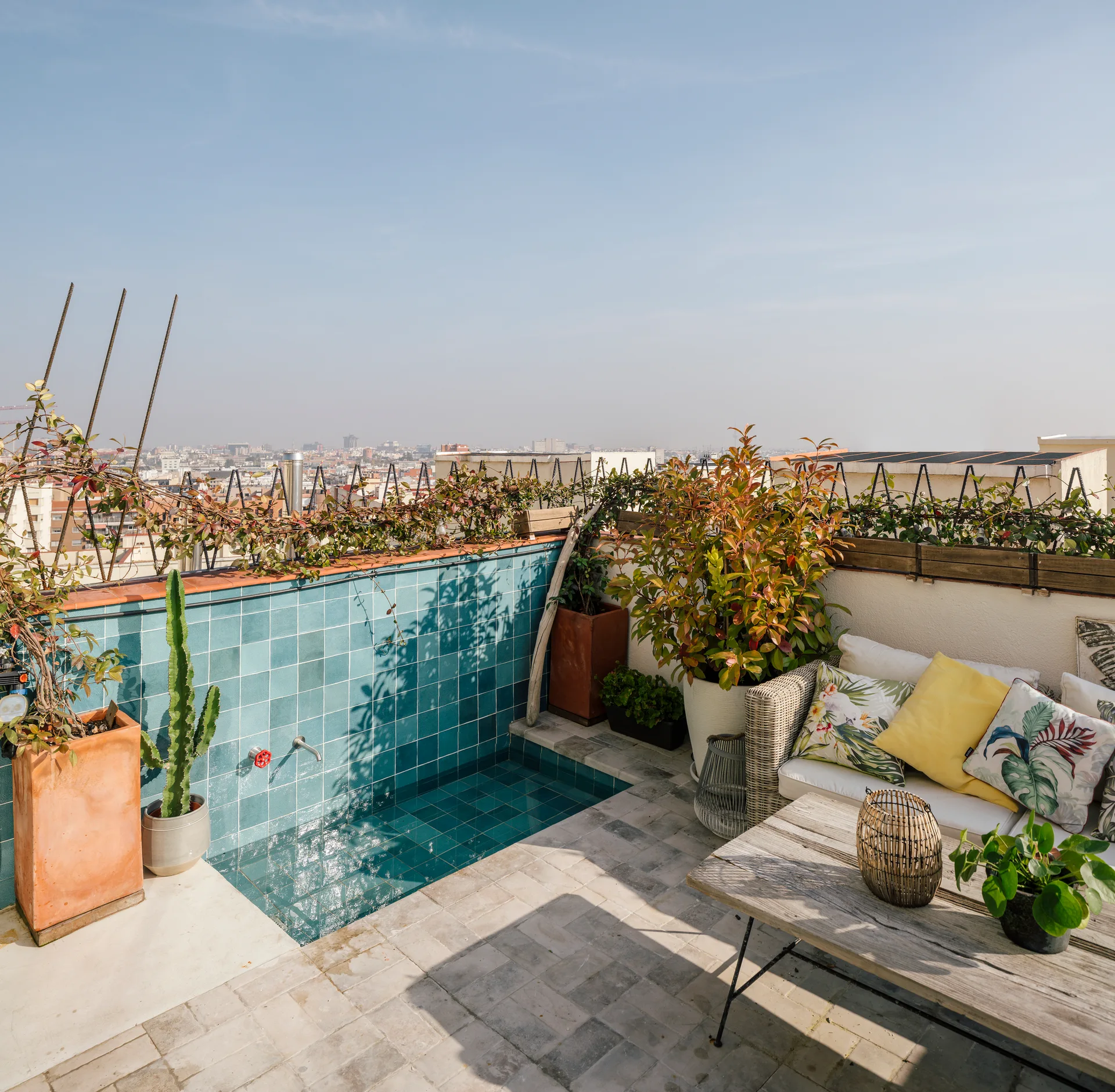 A sunny rooftop terrace features a small sunken pool lined with teal ceramic tiles next to an outdoor seating area with potted plants.