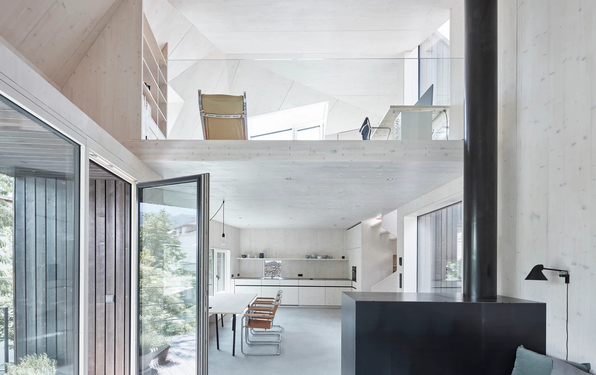 A bright, open-plan minimalist room features light wood paneling on walls and ceiling, a mezzanine loft with a glass railing, and a black fireplace column.