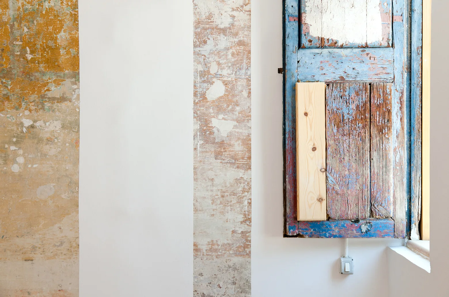 Alternating sections of smooth white plaster and distressed ochre paint next to a weathered blue wooden shutter on a wall.