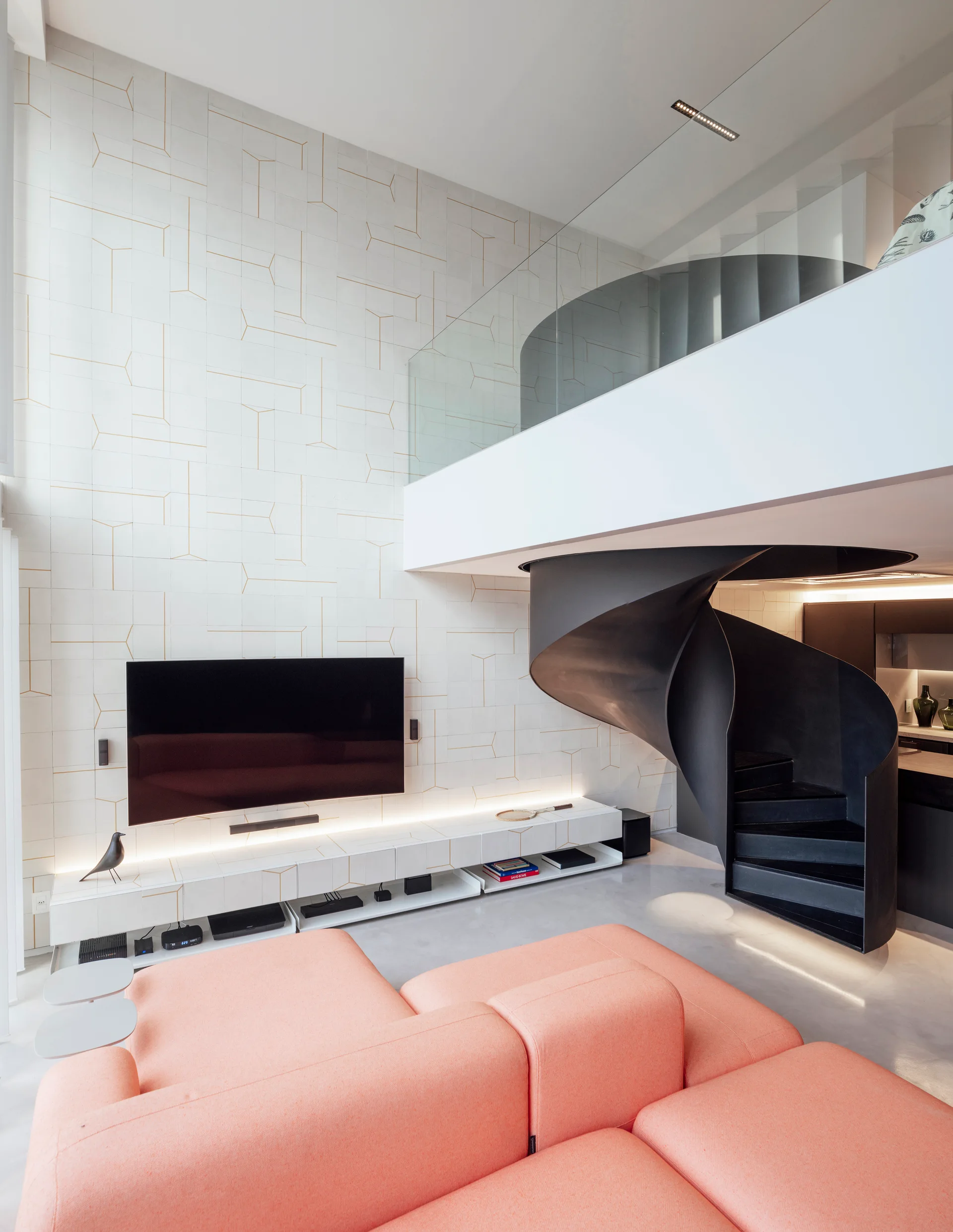 A modern living room features a peach modular sofa, a sculptural black spiral staircase, and a wall-mounted TV on a geometric tiled wall.