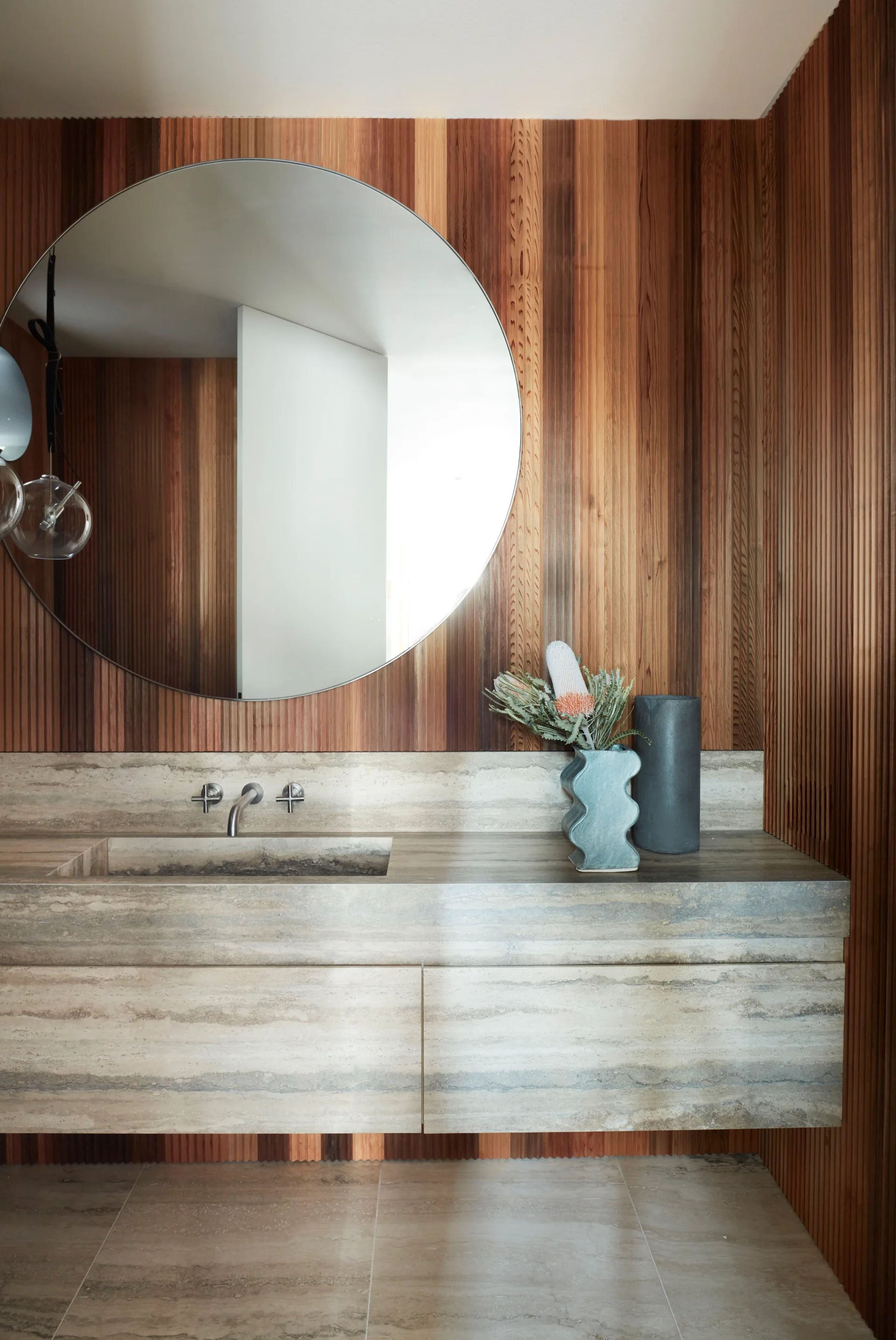 A floating travertine vanity with an integrated sink sits against a warm, vertical wood-slat wall beneath a large round mirror.