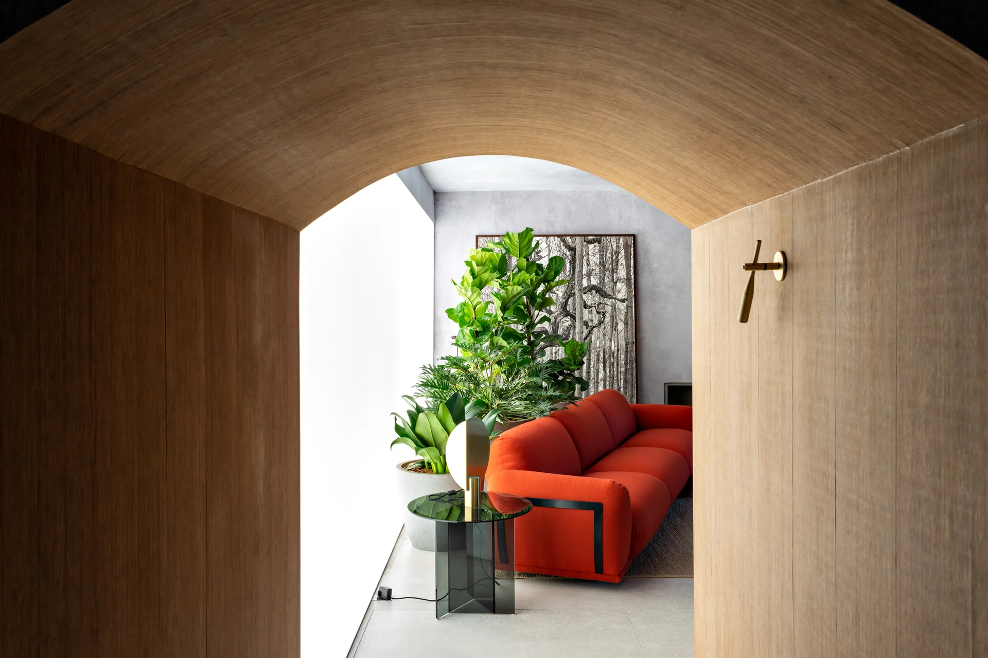 A view through a smooth wood-paneled archway into a bright living room featuring a bold red sofa, a large potted plant, and a glass side table.