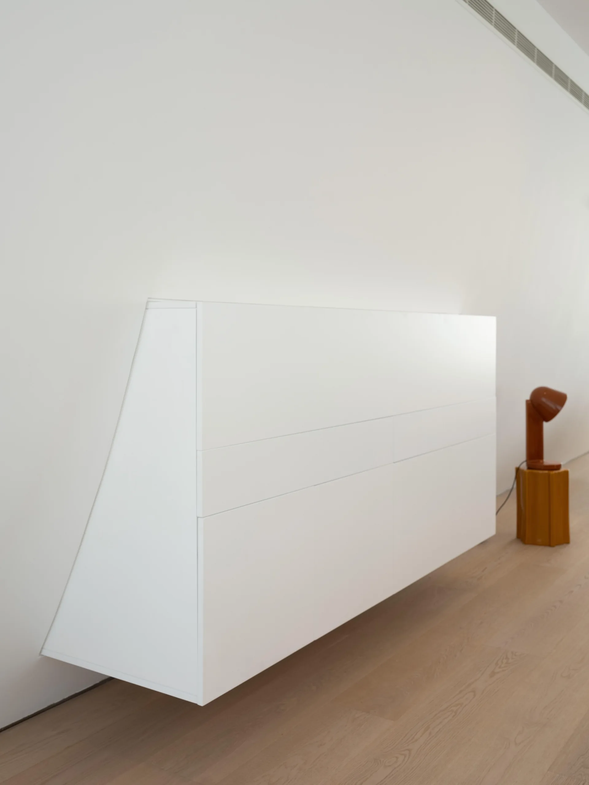 A minimalist white wall-mounted cabinet floats above light wood floors next to a small orange lamp on a pedestal.