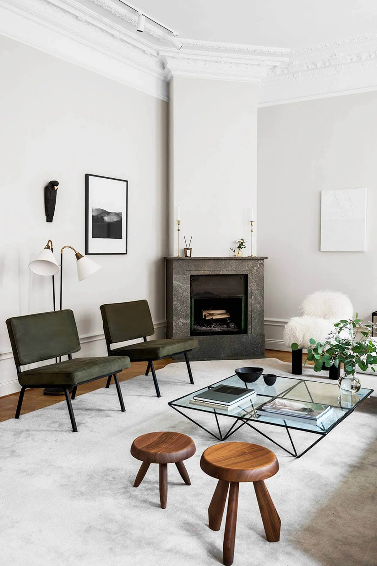 A bright living room features two olive green velvet armchairs, a geometric glass coffee table, and a grey stone fireplace.