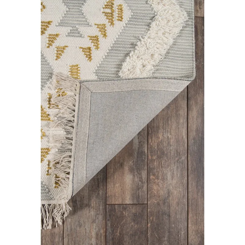 A corner of a grey and white geometric patterned rug with mustard yellow accents and fringe is folded back on a dark wood floor.