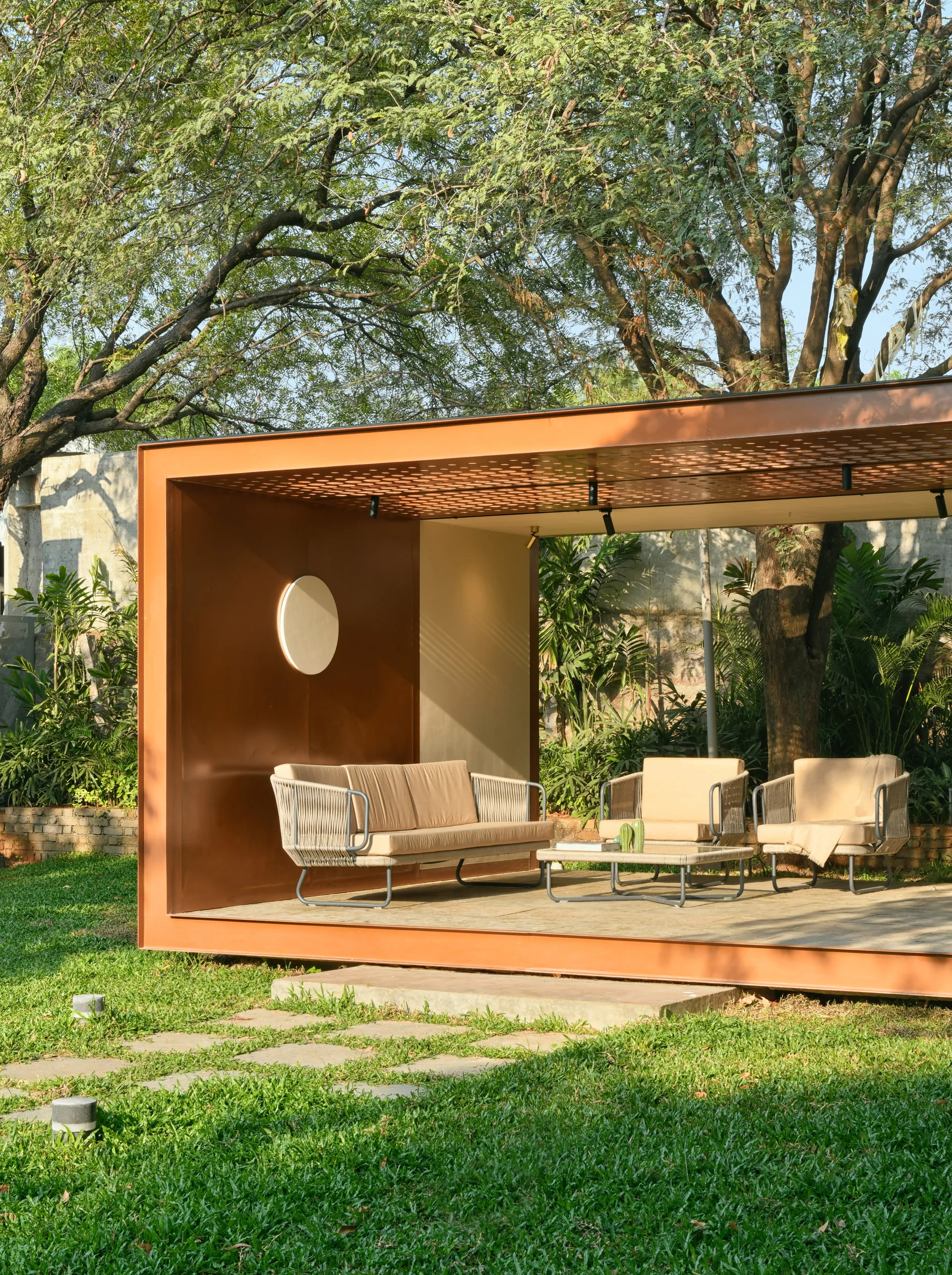 A modern rust-colored metal pavilion set on a green lawn features a beige outdoor sofa, matching woven armchairs, and a low coffee table.