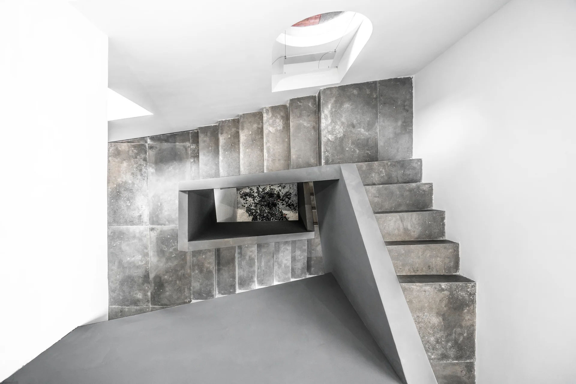 A high-angle view looking down a minimalist grey concrete staircase with white walls, a geometric handrail, and a circular skylight.