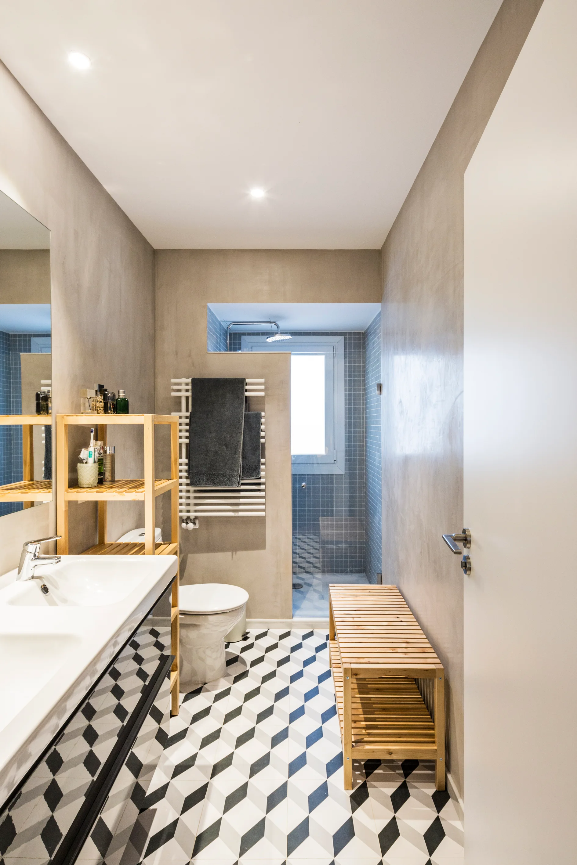 A contemporary bathroom features a black and white geometric tiled floor, smooth grey plaster walls, a double sink vanity, and light wood storage furniture.