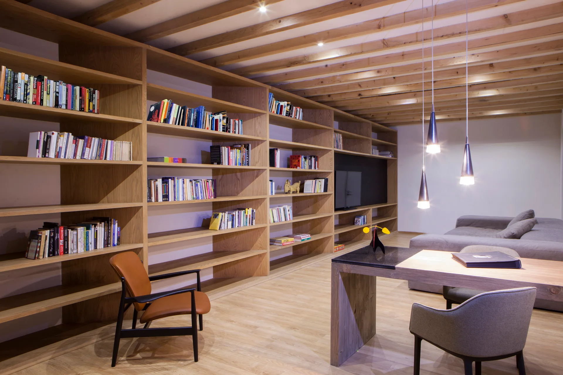 A home library features extensive floor-to-ceiling wooden bookshelves, exposed ceiling beams, a modern desk, and hanging pendant lights.