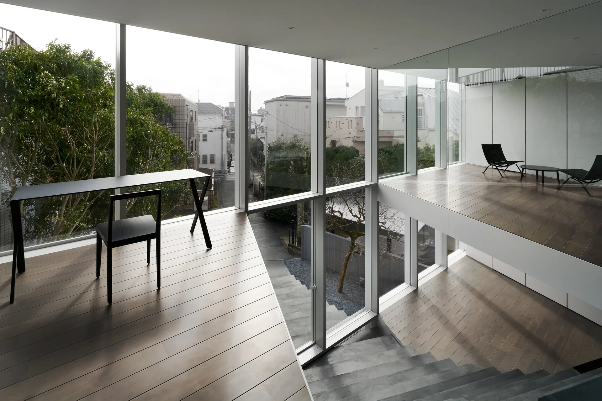 A modern multi level interior featuring wood floors, a grey concrete staircase, large glass walls, and a minimalist black desk with a chair.