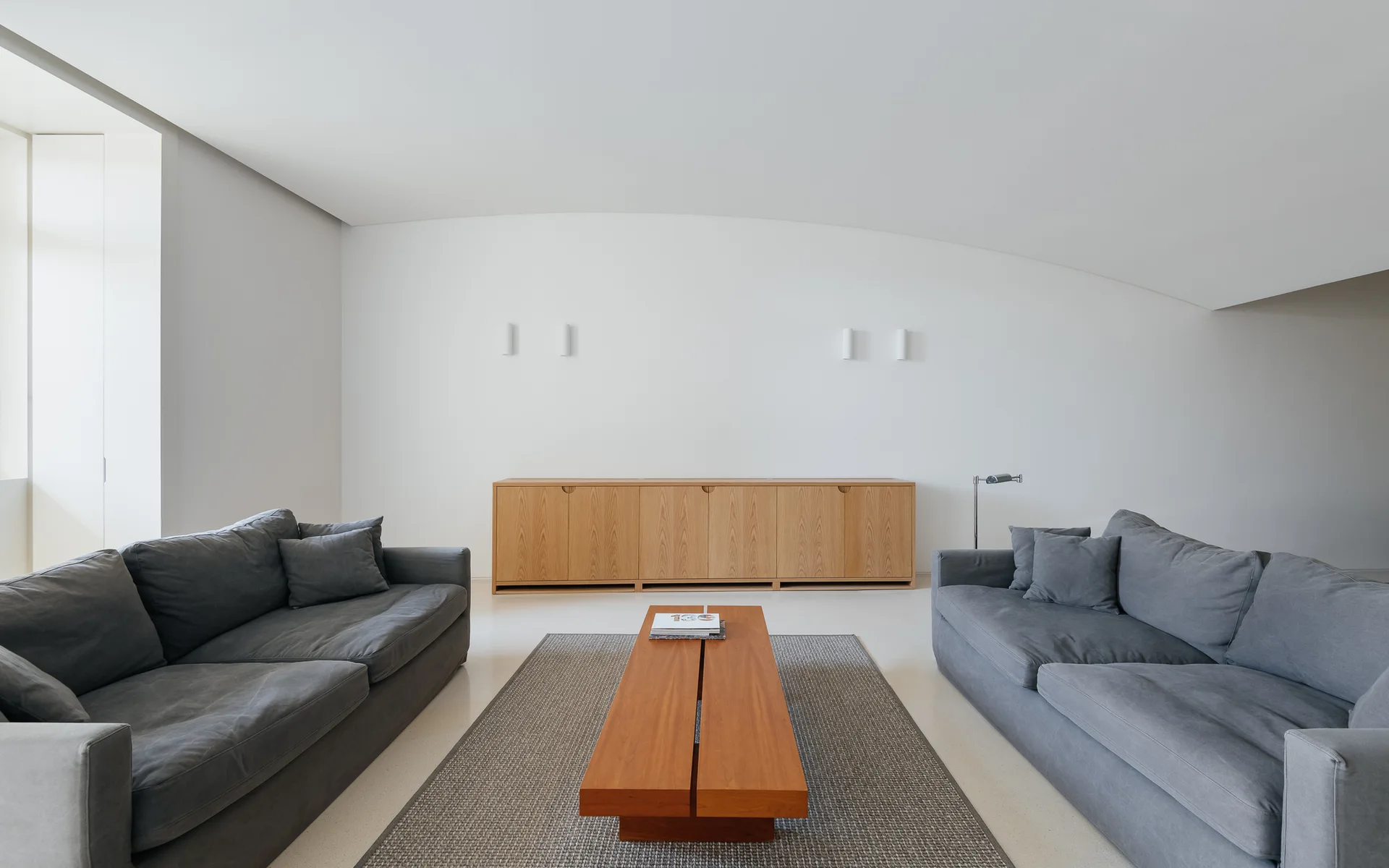 Two gray sofas face each other over a wooden coffee table and gray rug in a minimalist white living room with a wood sideboard.