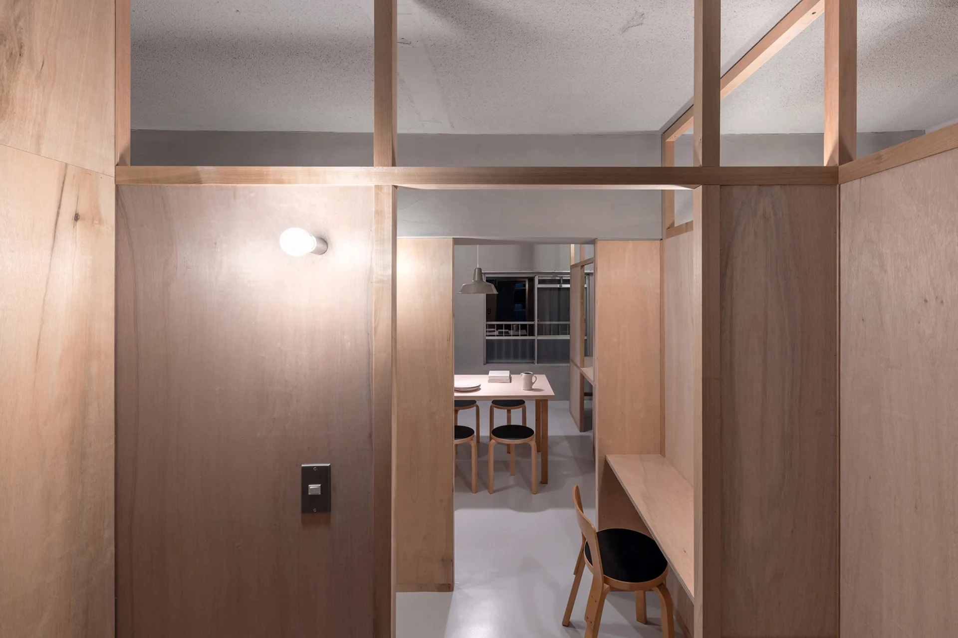 An open interior divided by raw plywood partitions, featuring a built-in study desk and a dining table with chairs in the background.