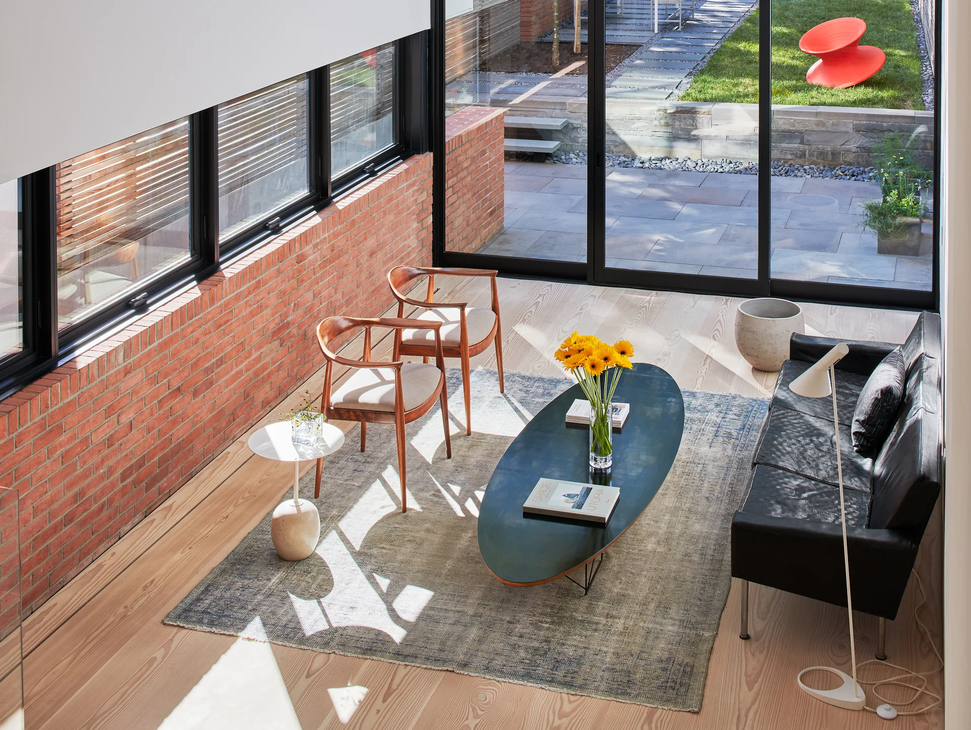 High-angle view of a living room with a black leather sofa, wooden armchairs, an exposed brick wall, and sliding glass doors opening to a garden.