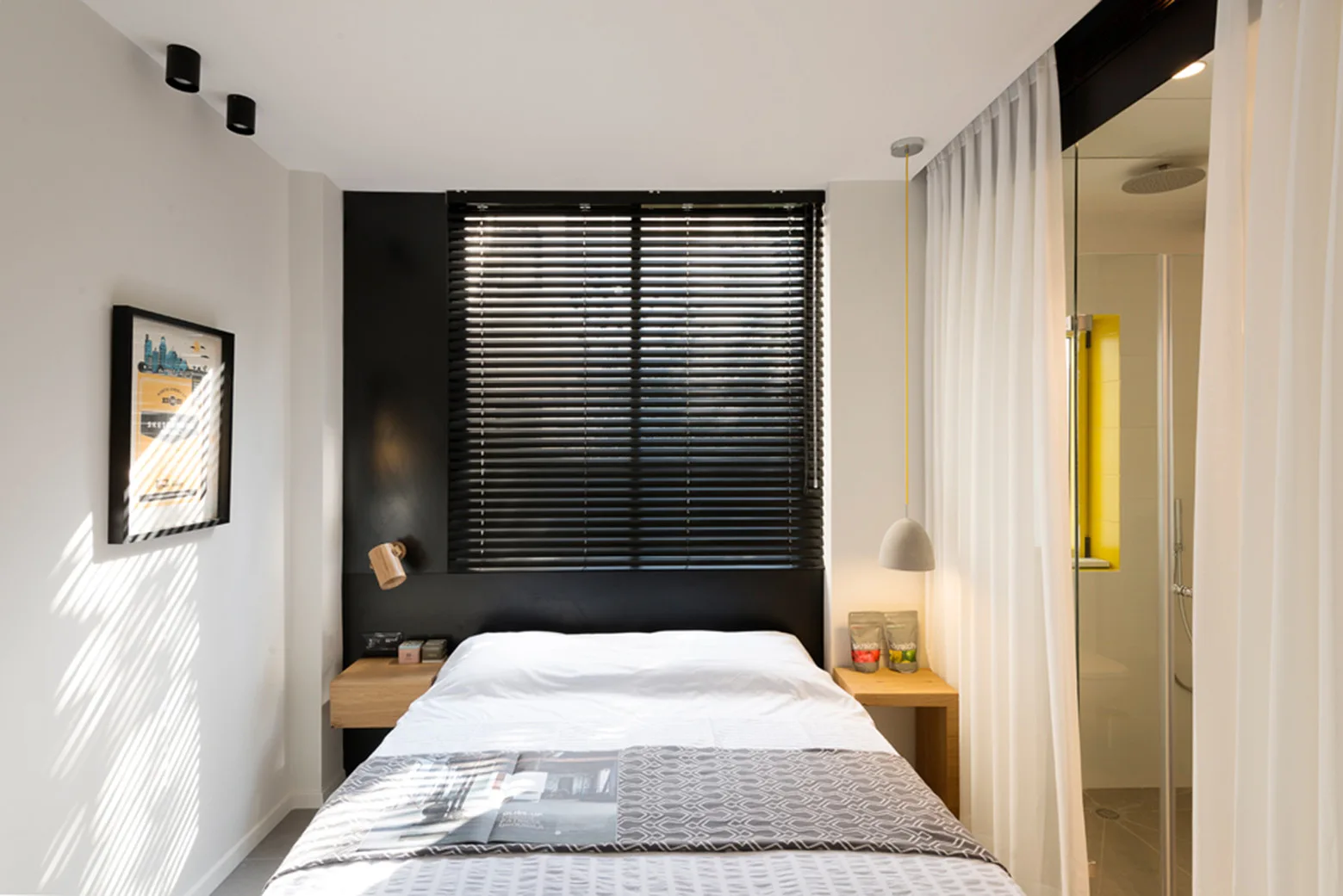 A modern bedroom features a bed with white linens, black window blinds, wooden nightstands, and an adjoining bathroom visible behind a sheer white curtain.