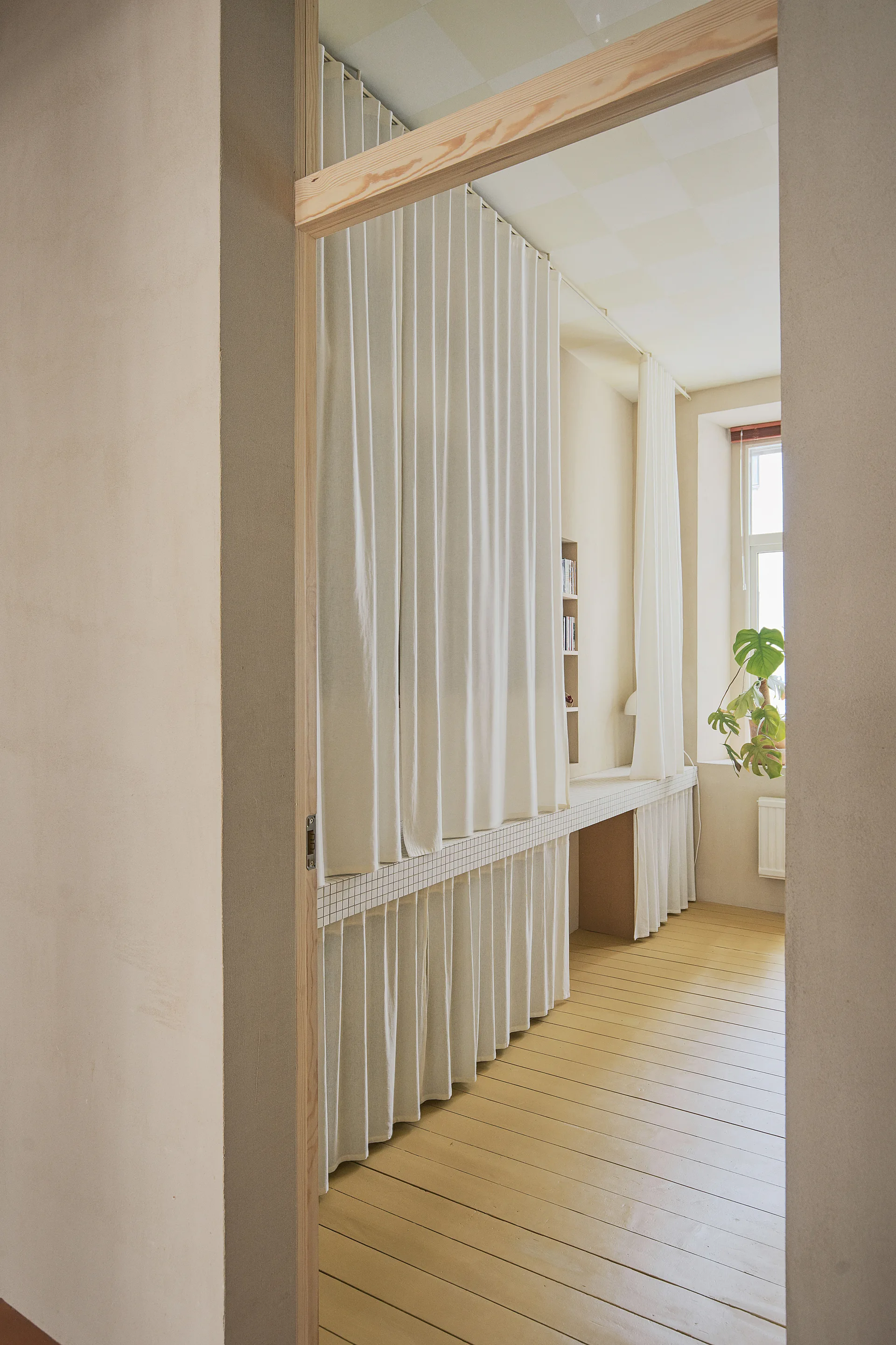 A minimalist cream room viewed through a wooden doorway features a long tiled counter concealed by gathered white fabric.