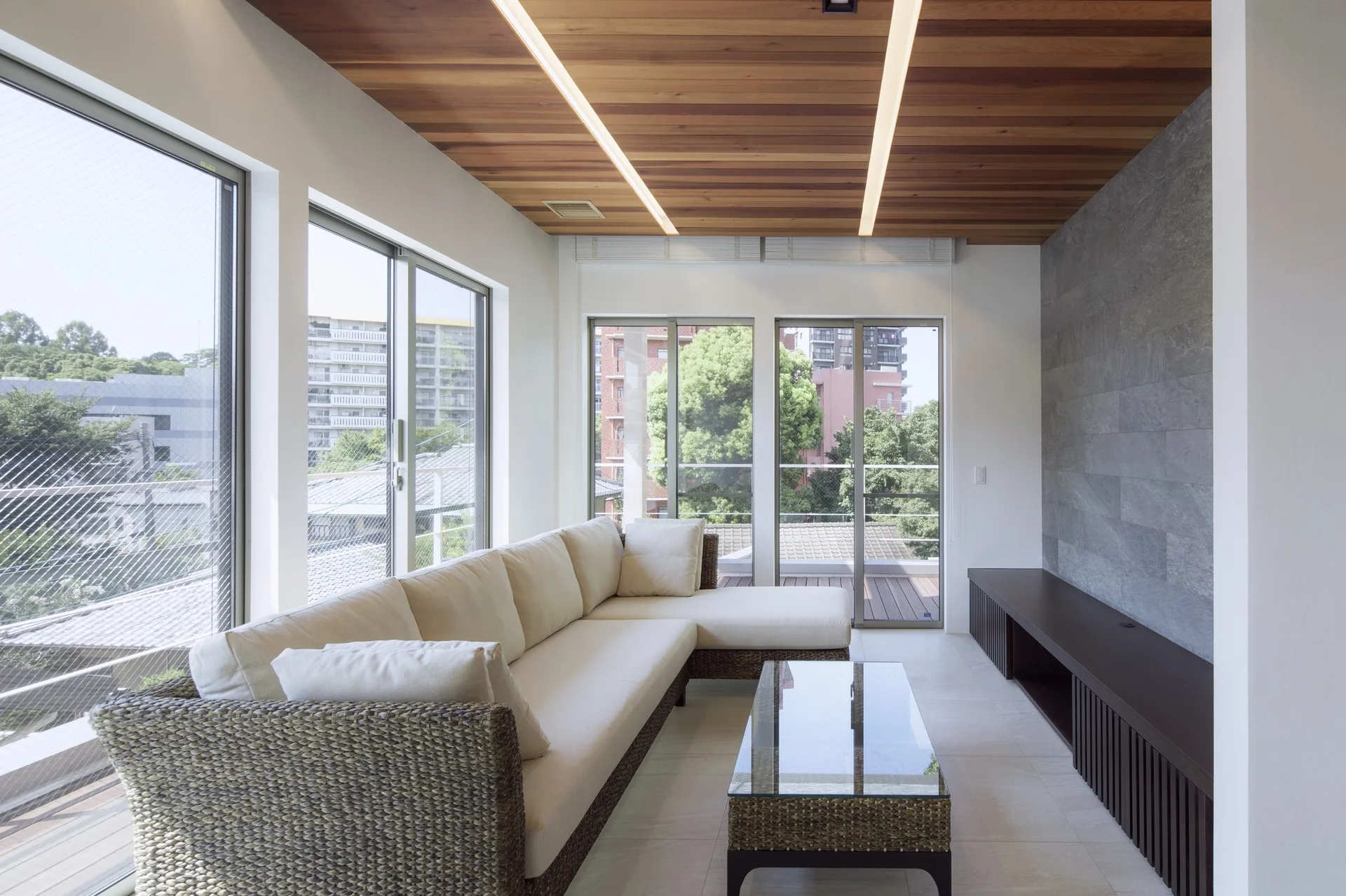 A modern living room featuring a cream sectional sofa with a woven wicker base, a wood plank ceiling with linear lights, and a grey stone accent wall.