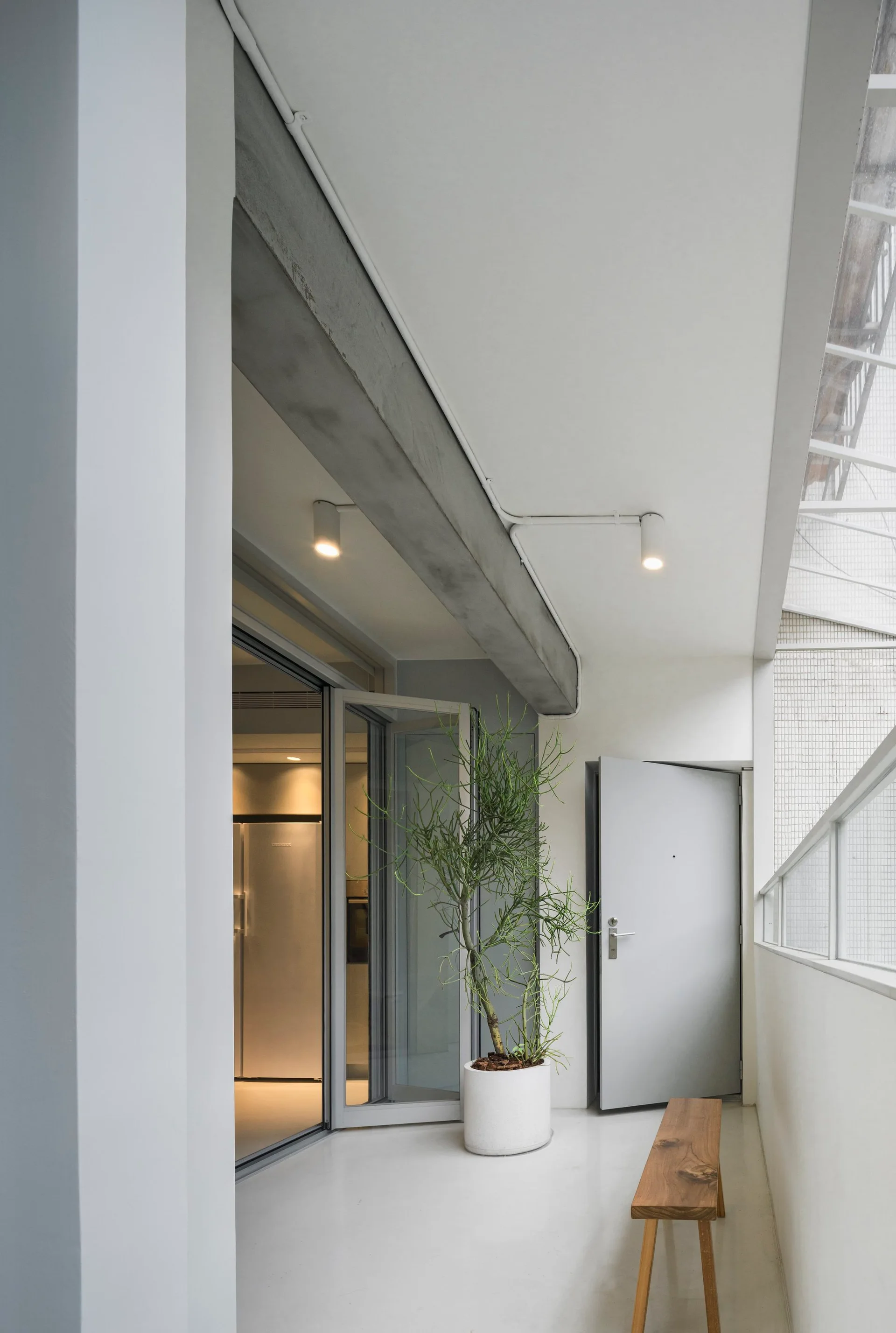 A minimalist balcony featuring a simple wooden bench, a potted plant, and a glass door under an exposed concrete ceiling beam.