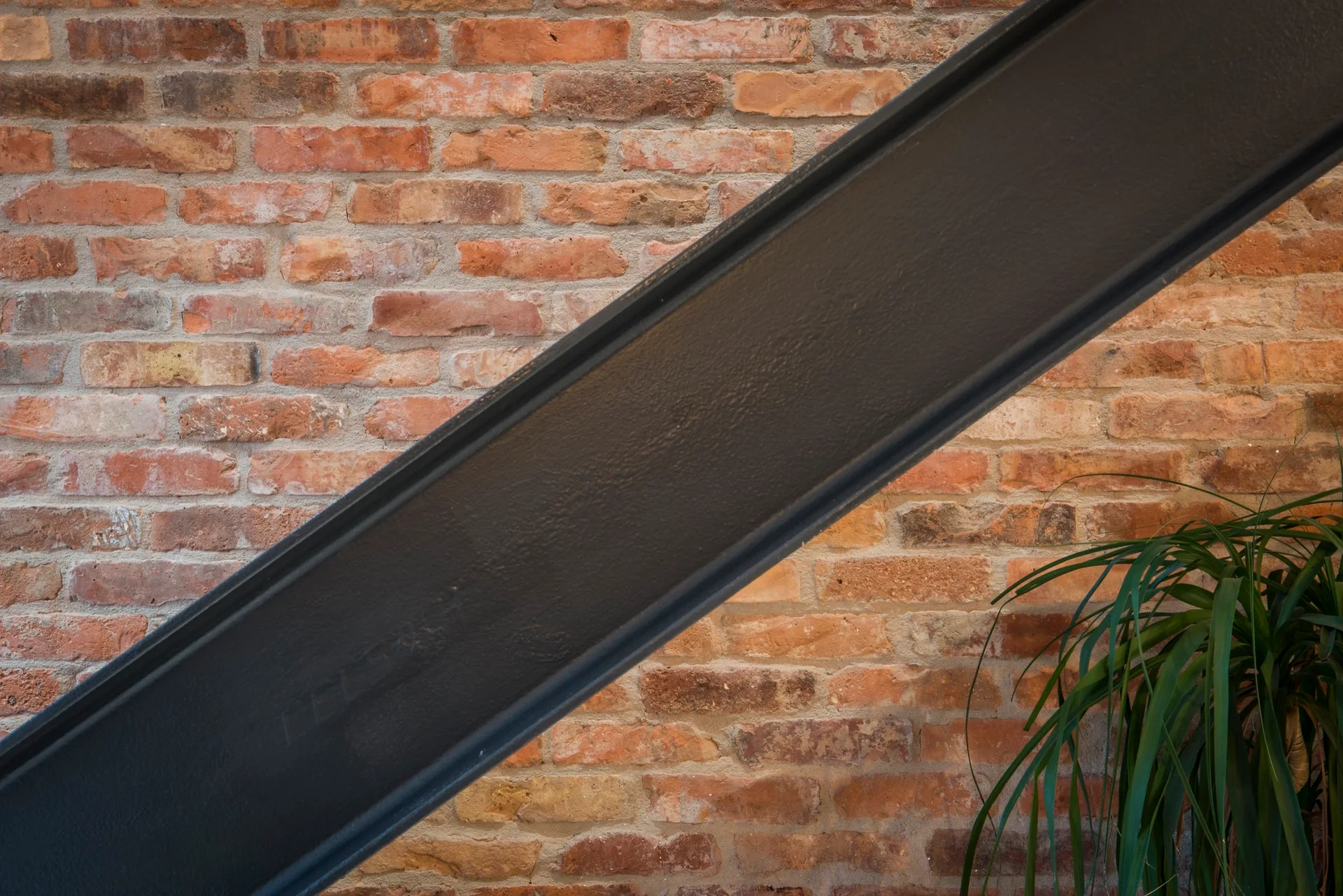 A dark metal structural beam runs diagonally across a textured red and brown exposed brick wall beside a green leafy plant.