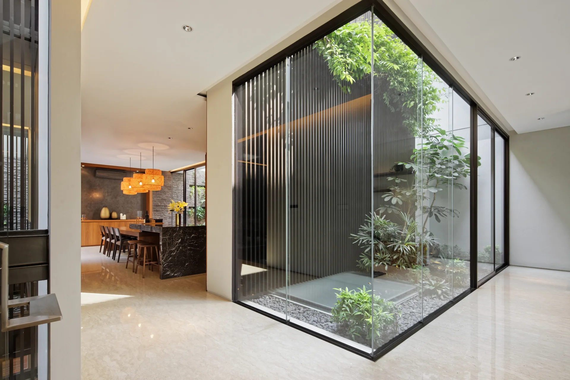 A modern open-plan interior featuring a glass-enclosed indoor courtyard with green plants next to a dining area with a dark marble island.