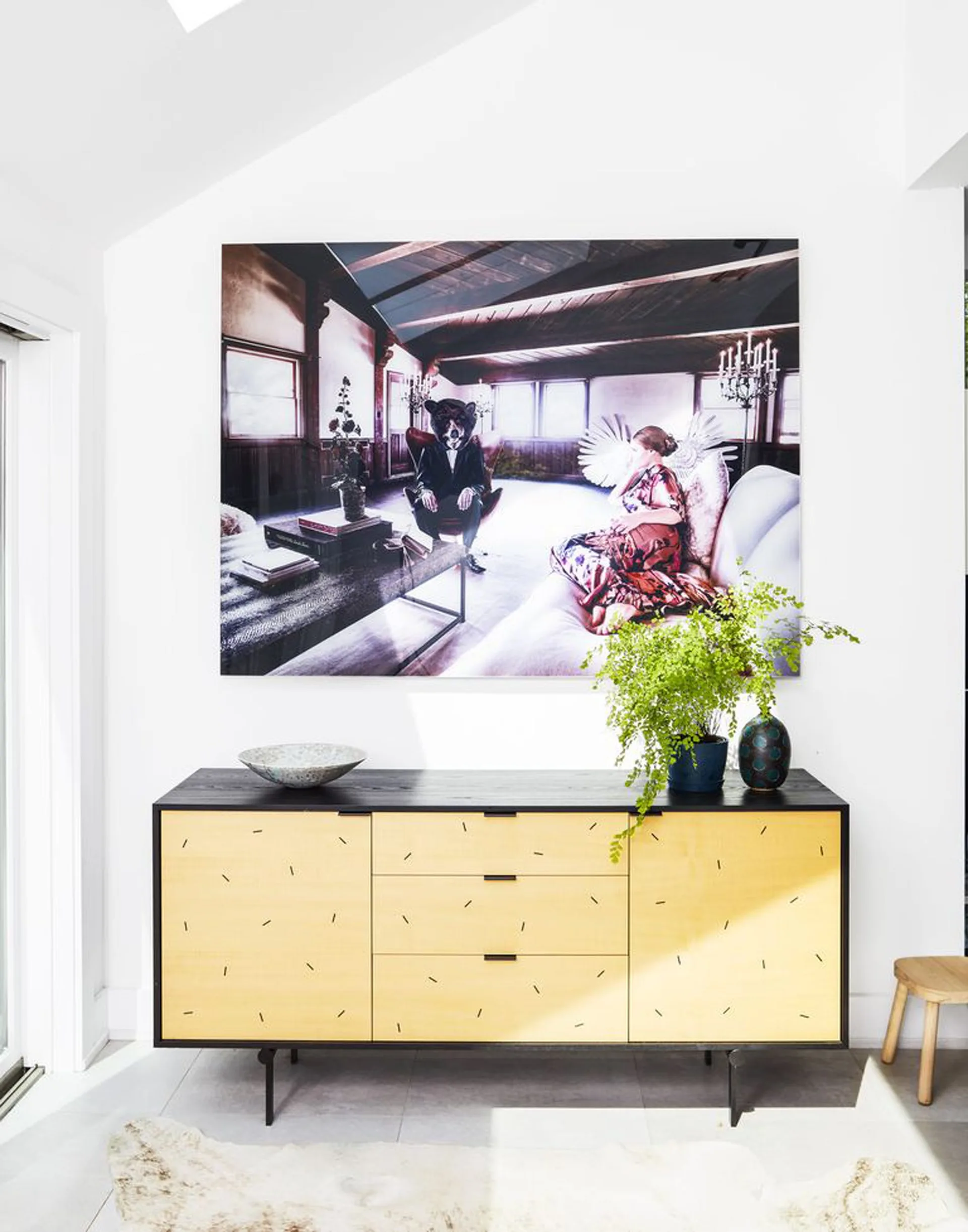 A modern yellow and black patterned credenza sits against a white wall beneath a large surreal art piece and a potted green fern.