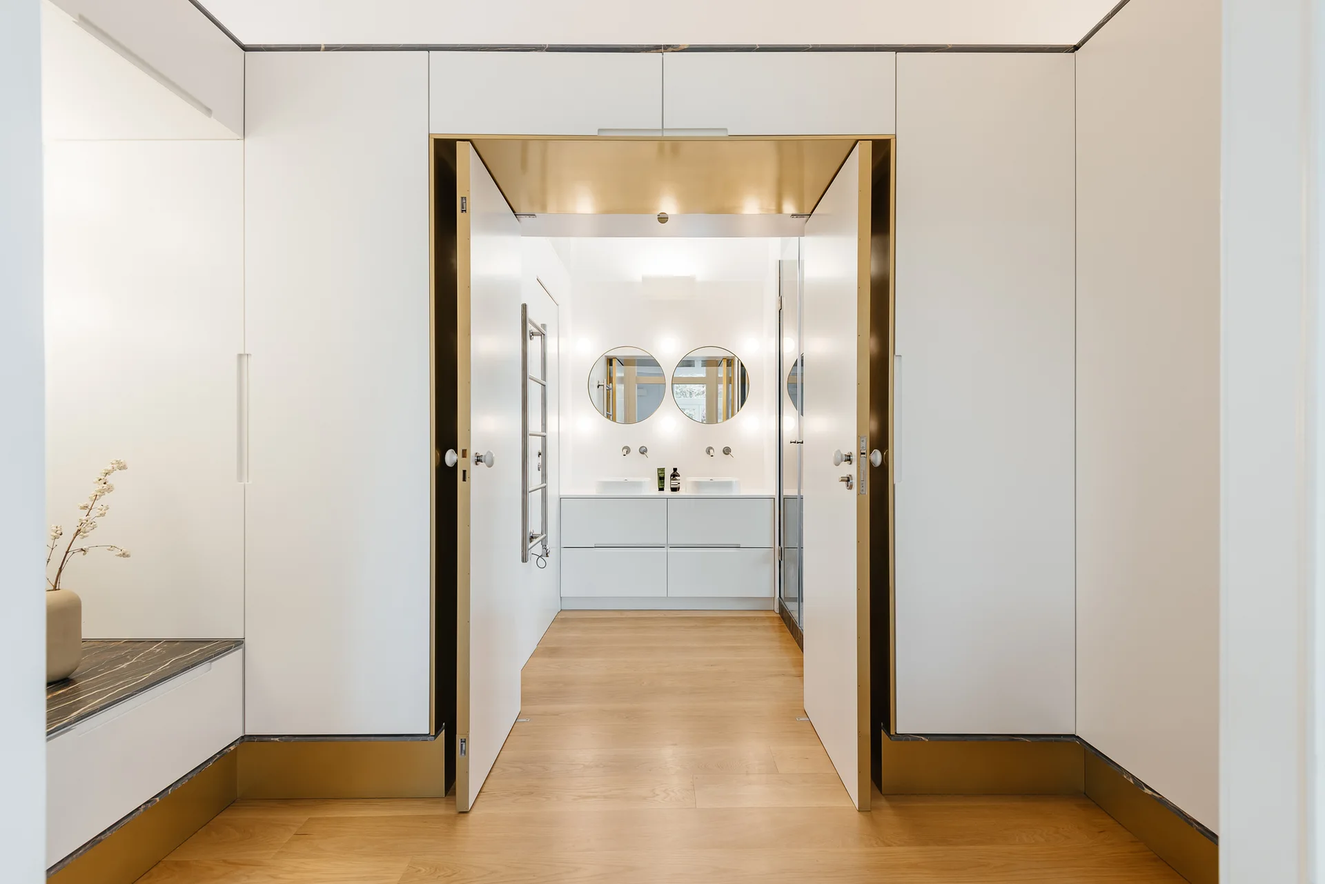 White double doors with a gold trim entryway open to a minimalist bathroom with a double vanity and two round mirrors.