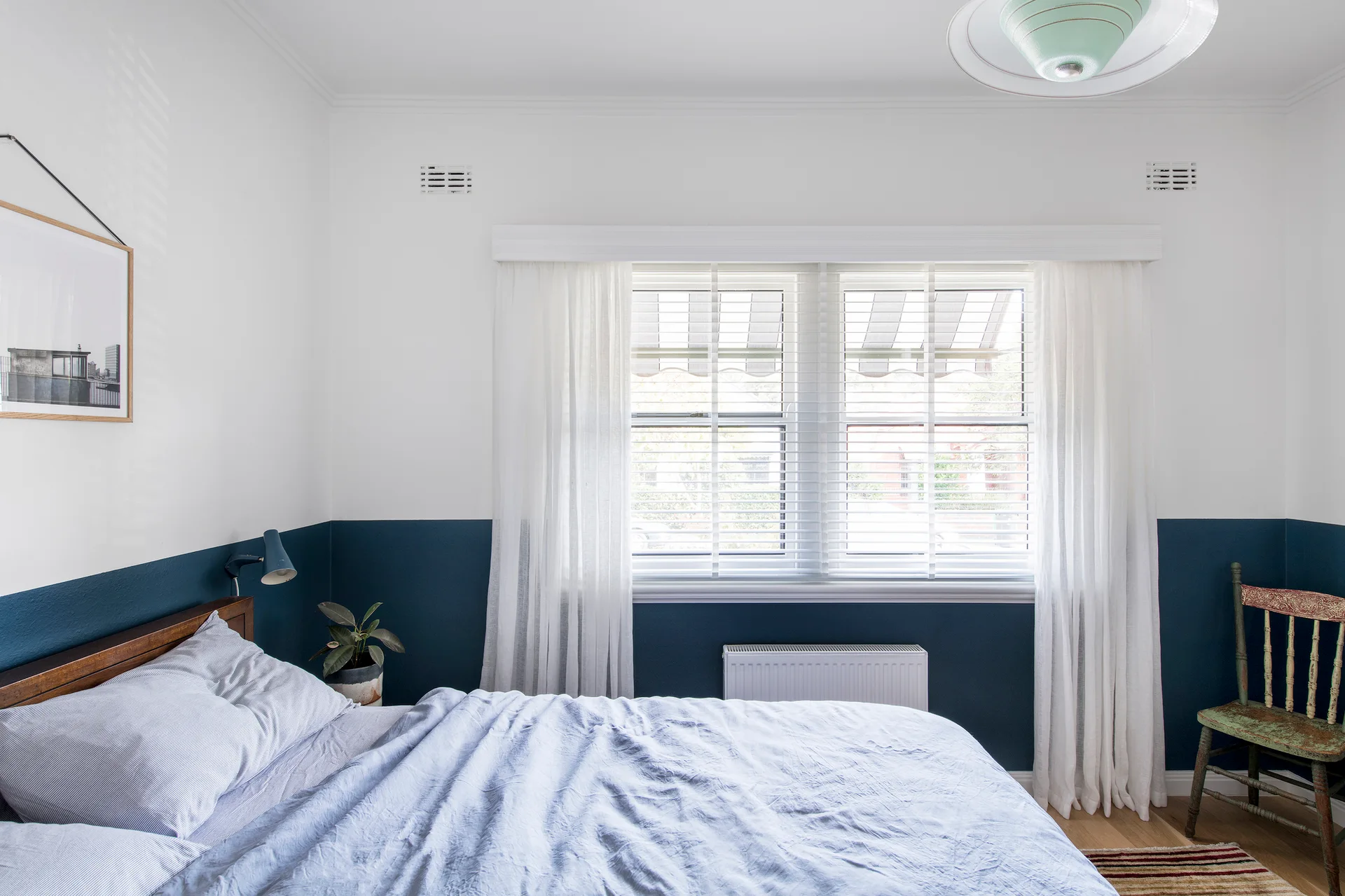 A bedroom features a bed with light blue bedding set against a two-tone navy and white wall near a window with sheer curtains.