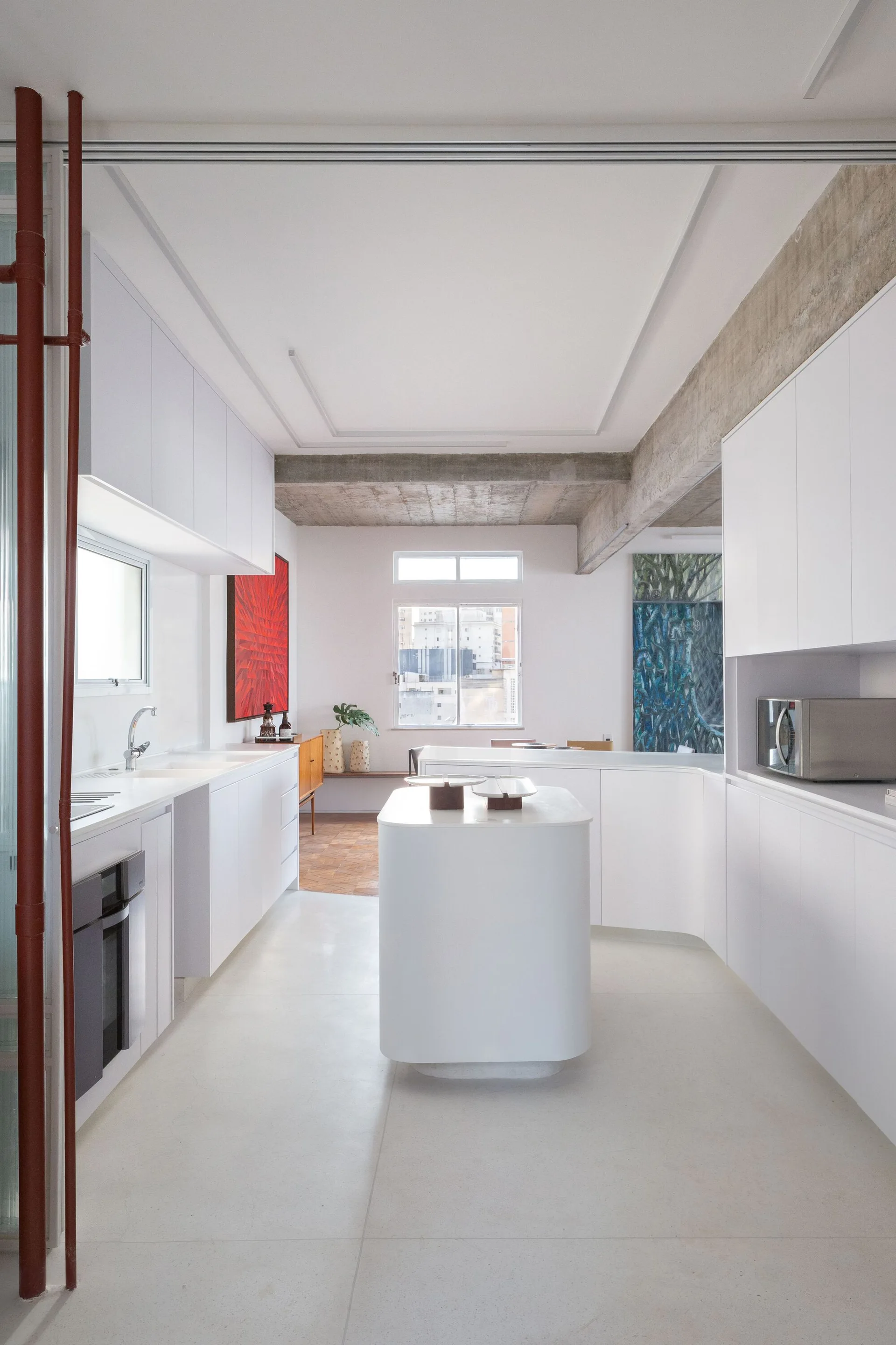 A bright minimalist kitchen features flat-panel white cabinetry, a curved central island, and an exposed raw concrete ceiling beam.