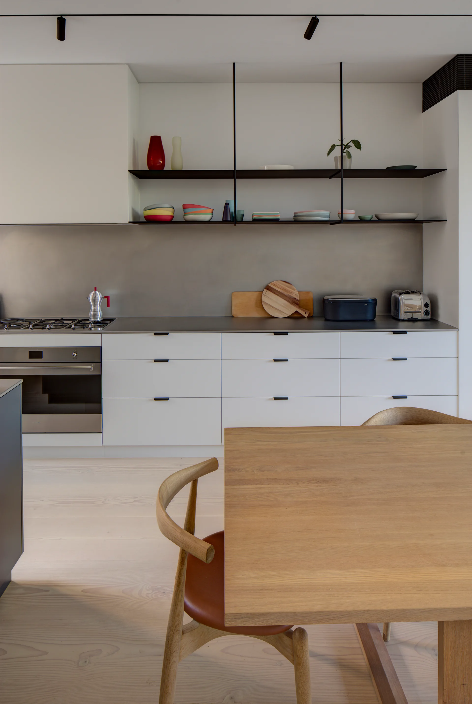A modern kitchen featuring white lower cabinetry, a brushed metal backsplash, open dark shelving, and a light wood dining table in the foreground.