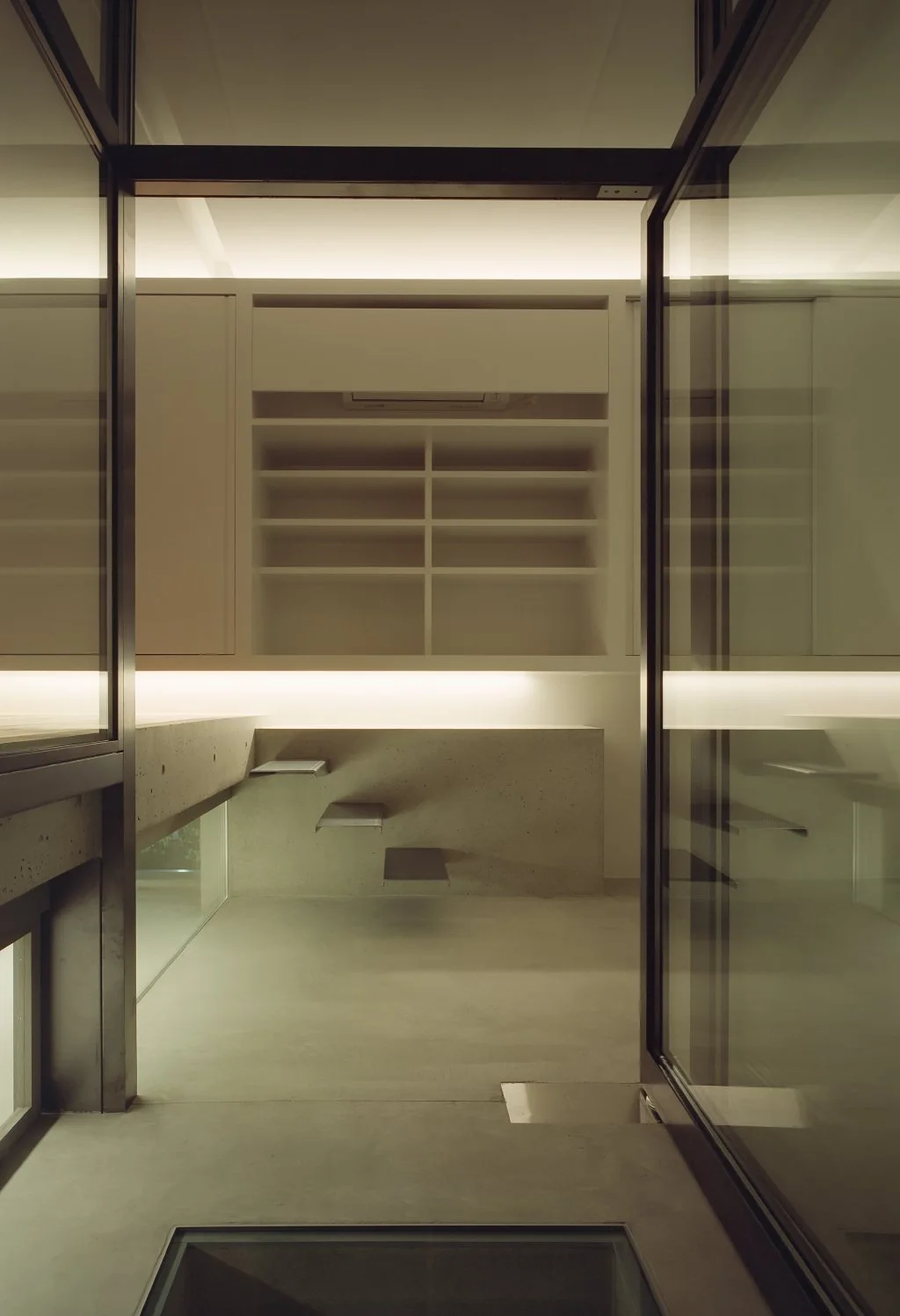A minimalist interior featuring a raw concrete wall with floating steps and white built-in shelving, viewed through a glass doorway.