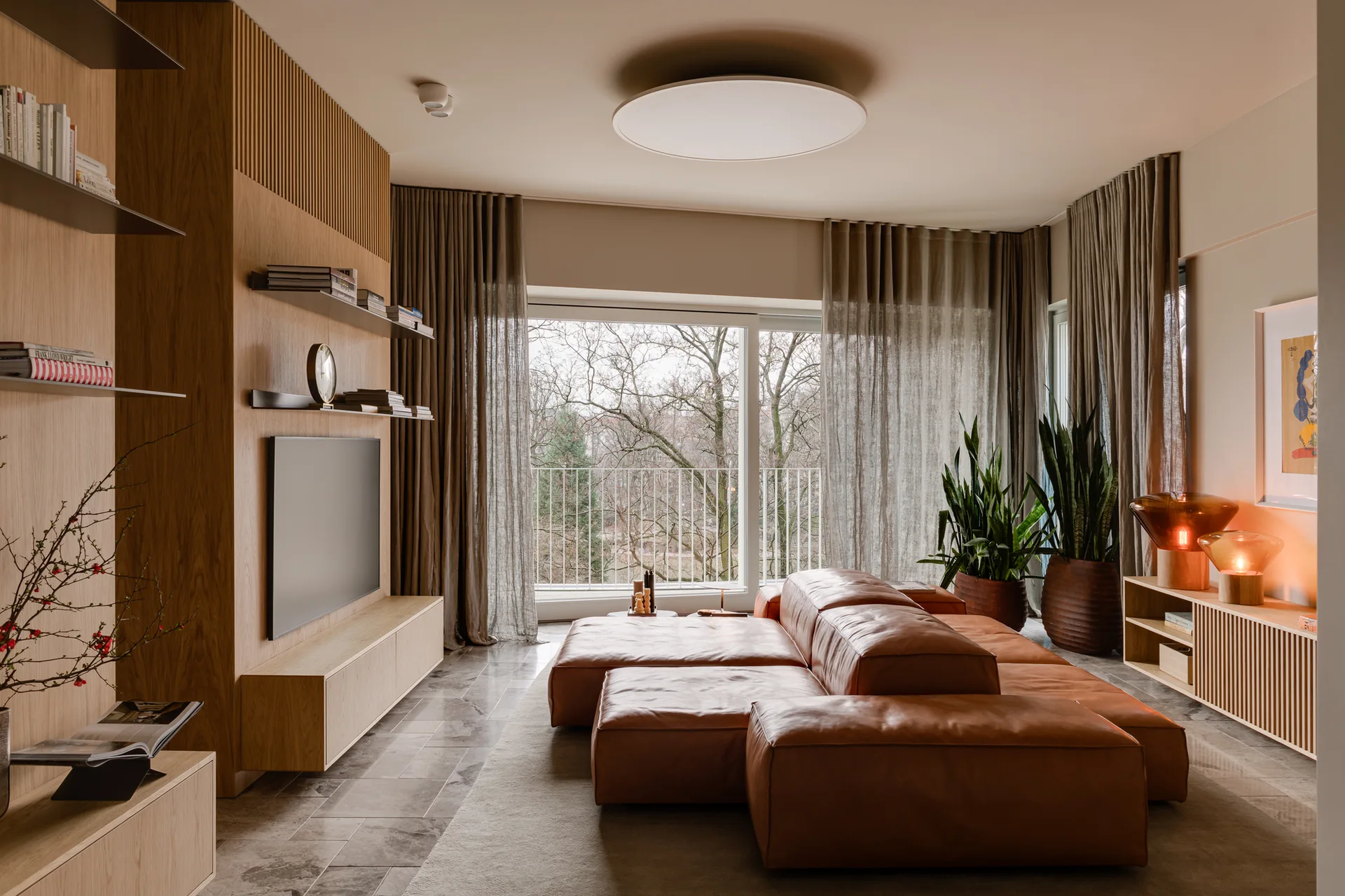 A contemporary living room featuring a modular tan leather sofa, a built-in wood media wall, and large windows with sheer drapes.