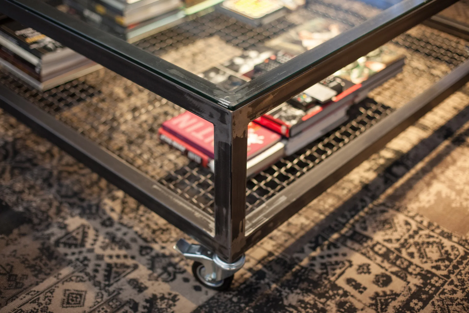 An industrial metal coffee table with a glass top and a mesh lower shelf containing books sits on a dark patterned rug.