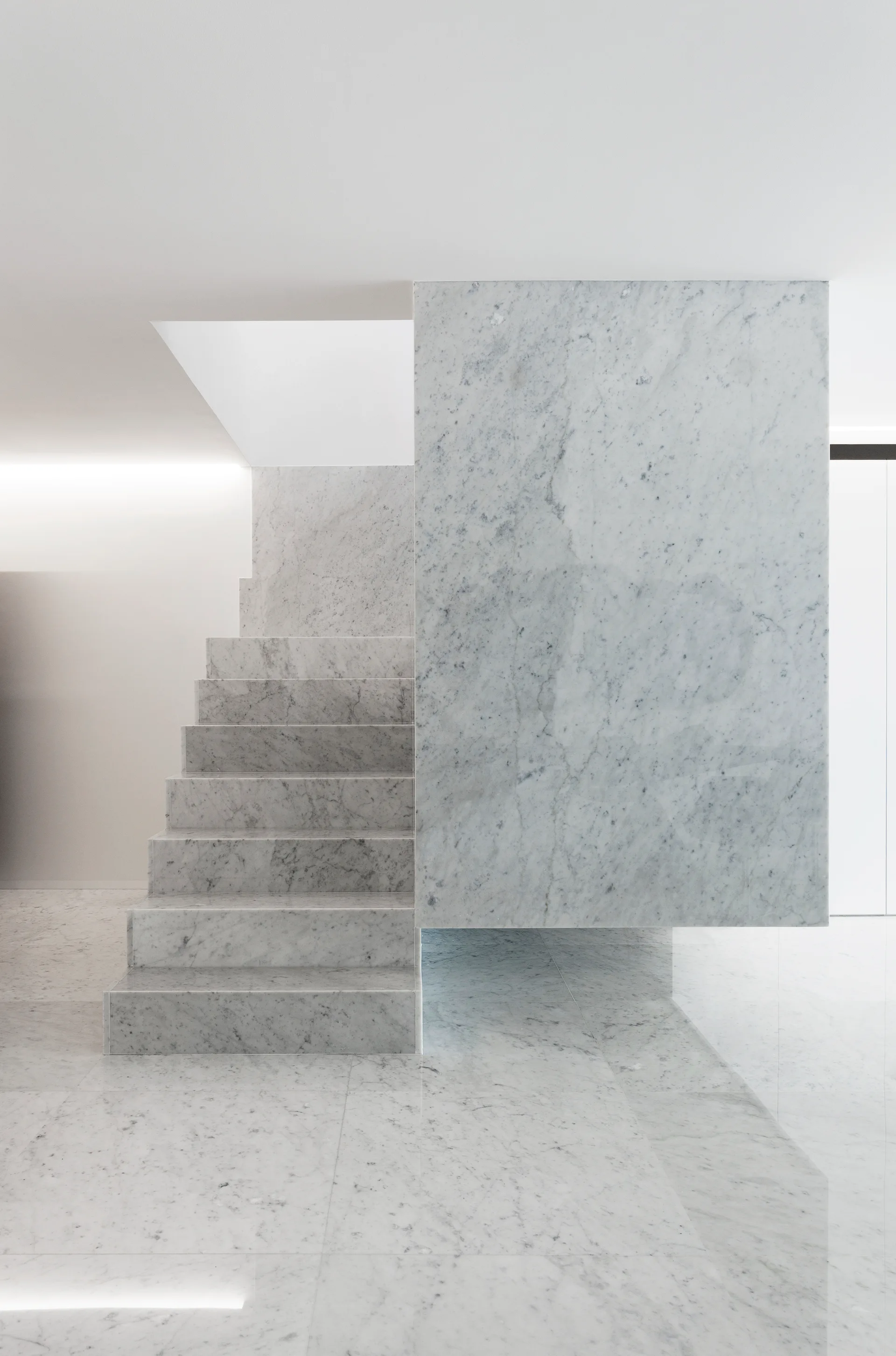 A minimalist staircase constructed from white veined marble stands on a matching polished floor next to a suspended stone partition wall.