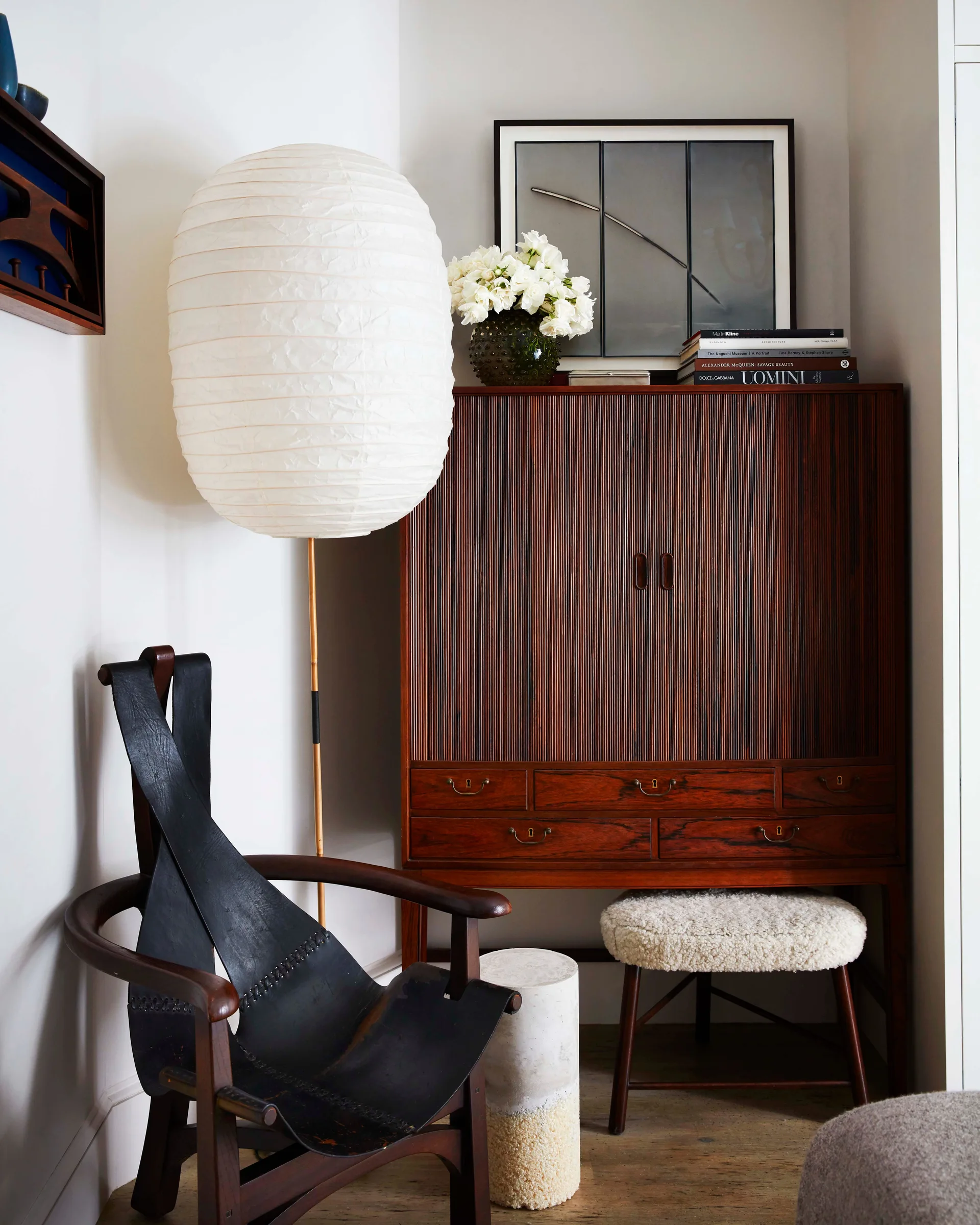 A cozy room corner featuring a dark fluted wood cabinet, a black leather strap armchair, a tall paper lantern lamp, and a small textured footstool.