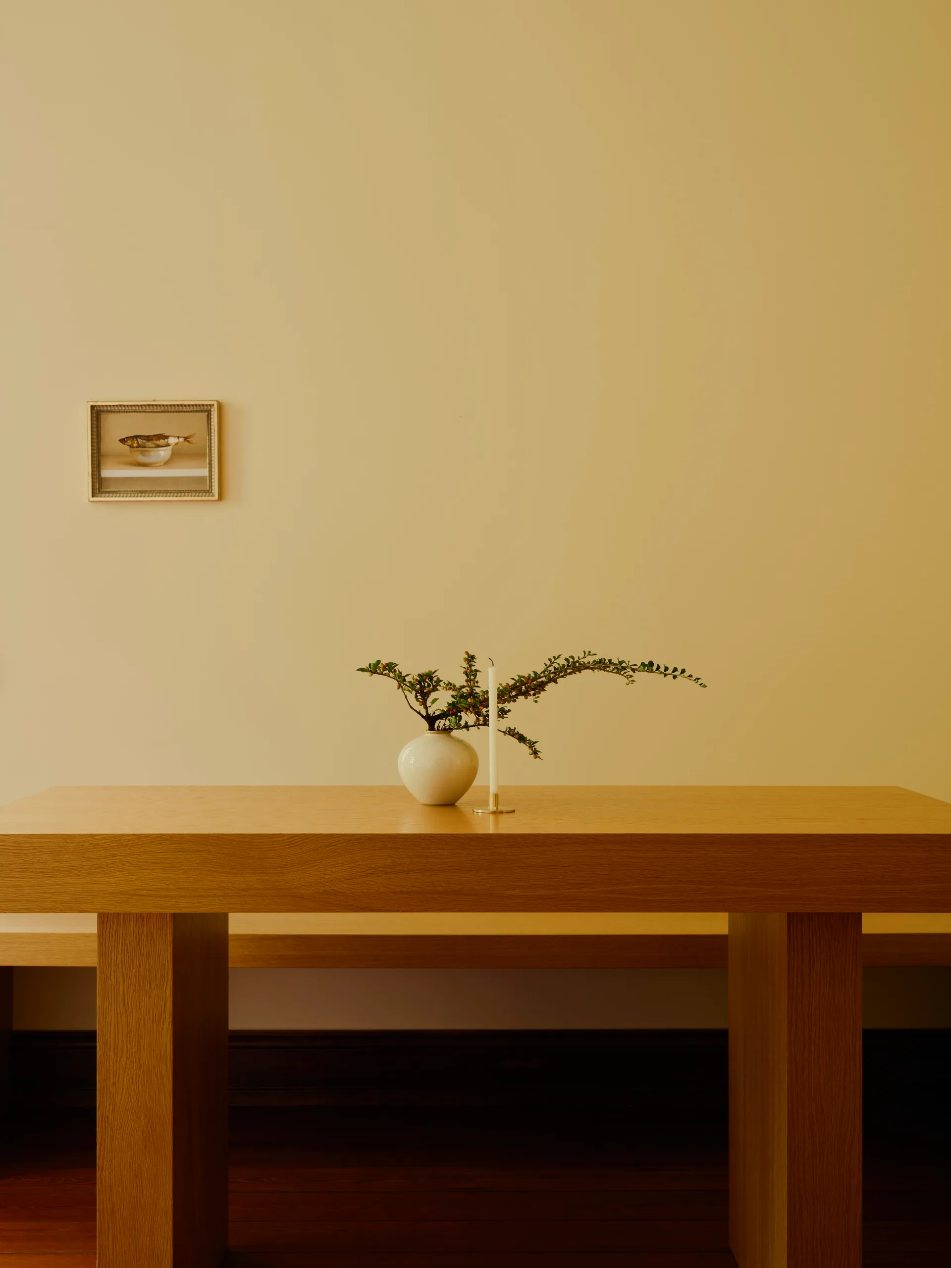 A wooden dining table features a white ceramic vase with a green branch and a single taper candle against a warm beige wall with a small framed picture.