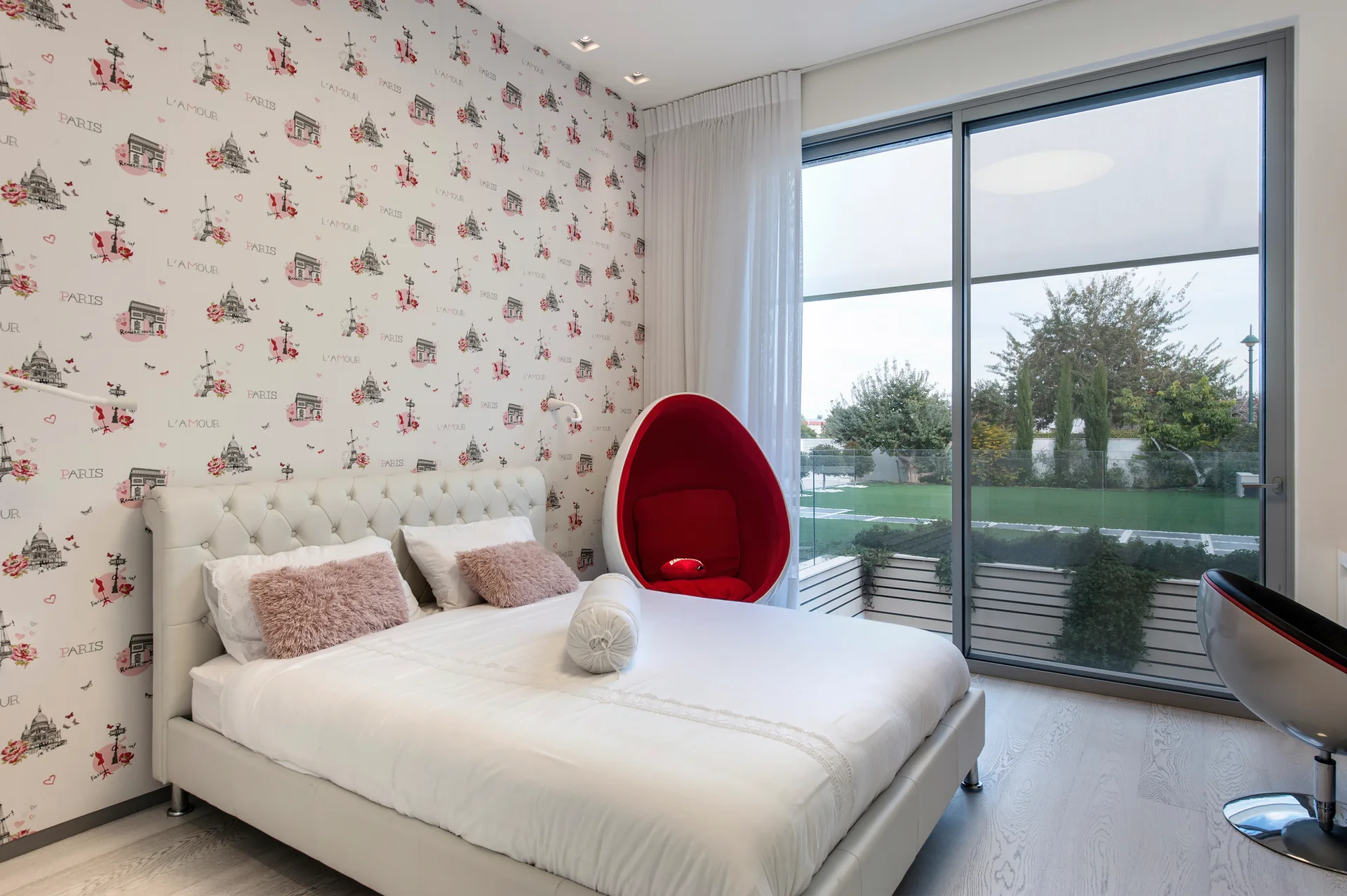 A modern bedroom features a tufted white leather bed, Paris-themed patterned wallpaper, and a red egg chair next to a large sliding glass door.