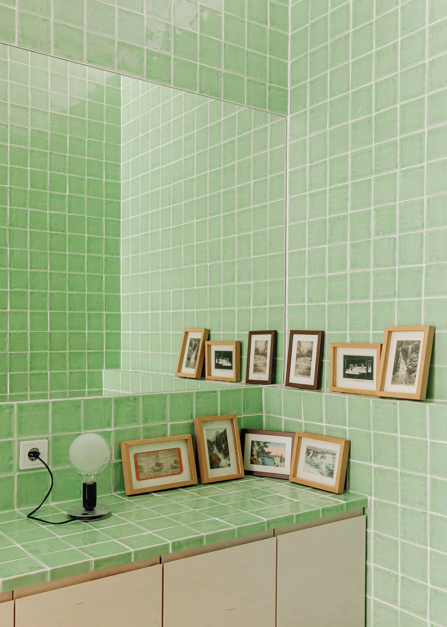 A bathroom vanity and wall covered in glossy green square tiles features several small wooden-framed landscape pictures leaning against a large mirror.