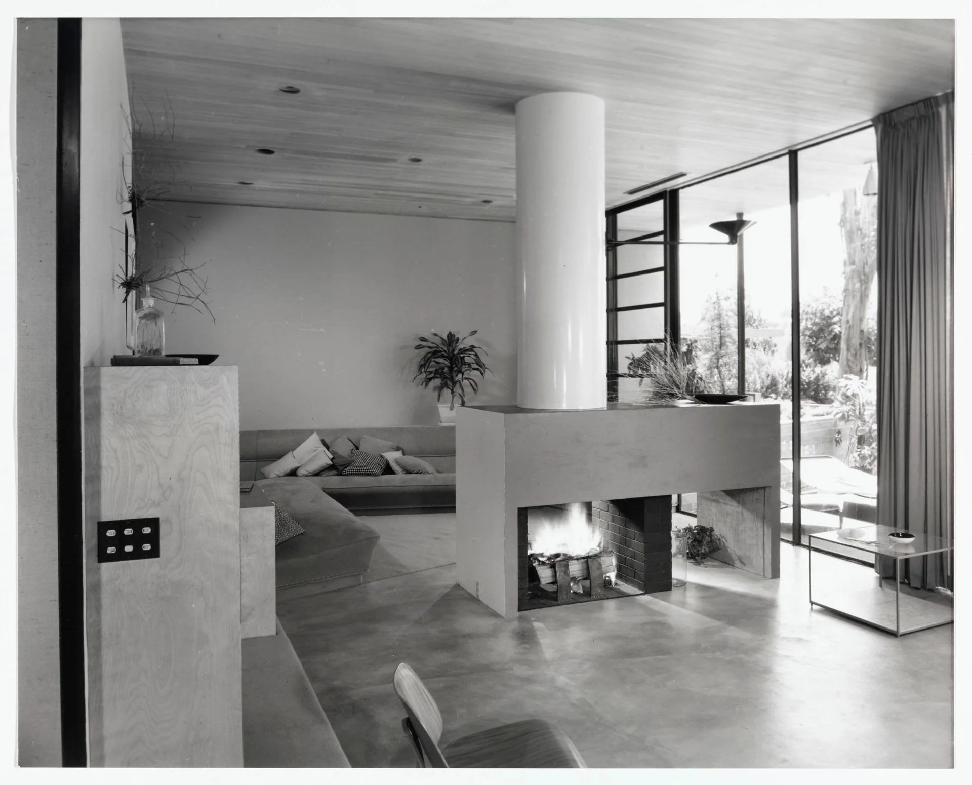 A mid-century modern living room features a central fireplace with a cylindrical white chimney, a wood-paneled ceiling, and glass walls.