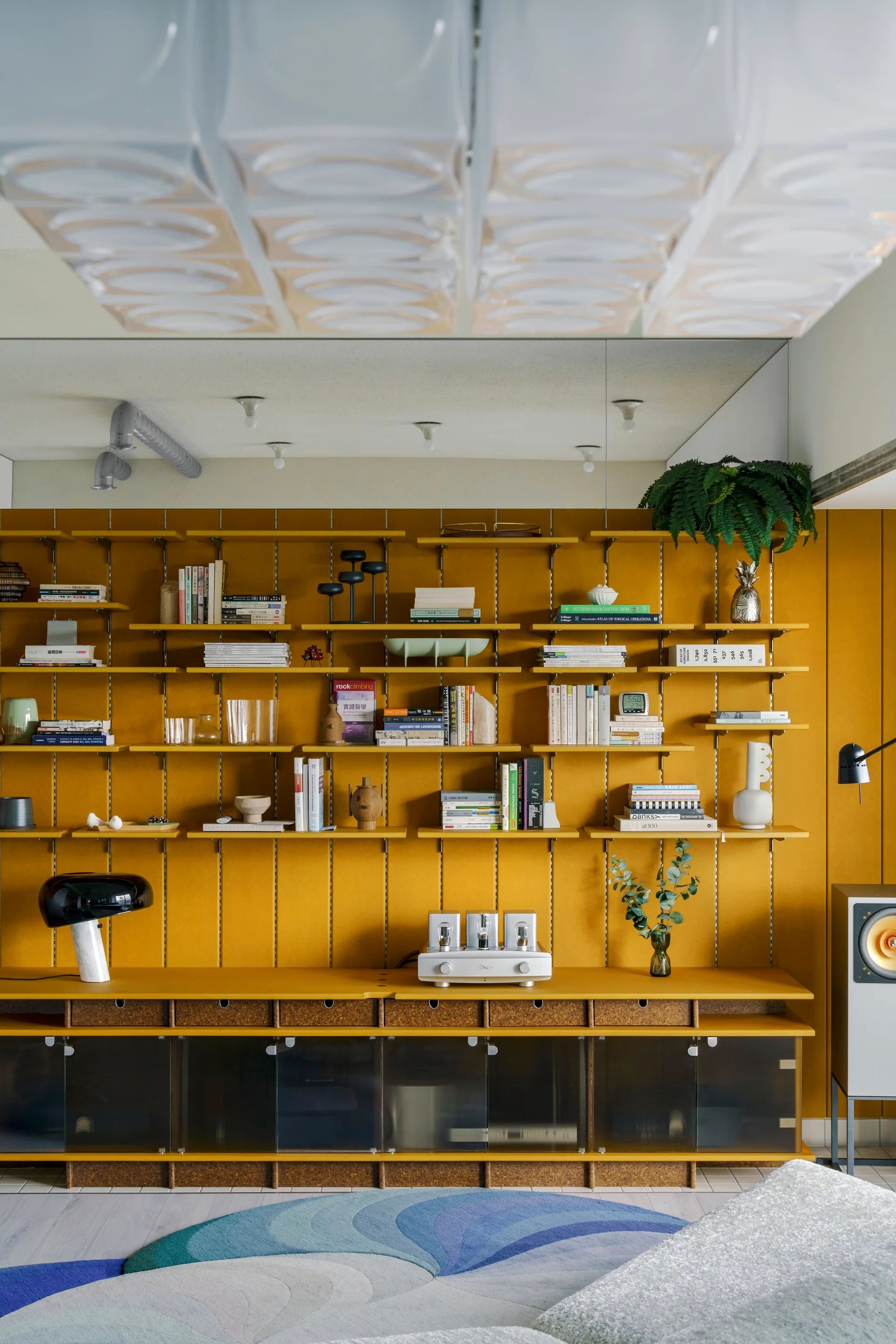 A wall-mounted mustard yellow modular shelving system sits against a fabric-paneled wall, holding books, decor, and audio equipment above glass cabinets.
