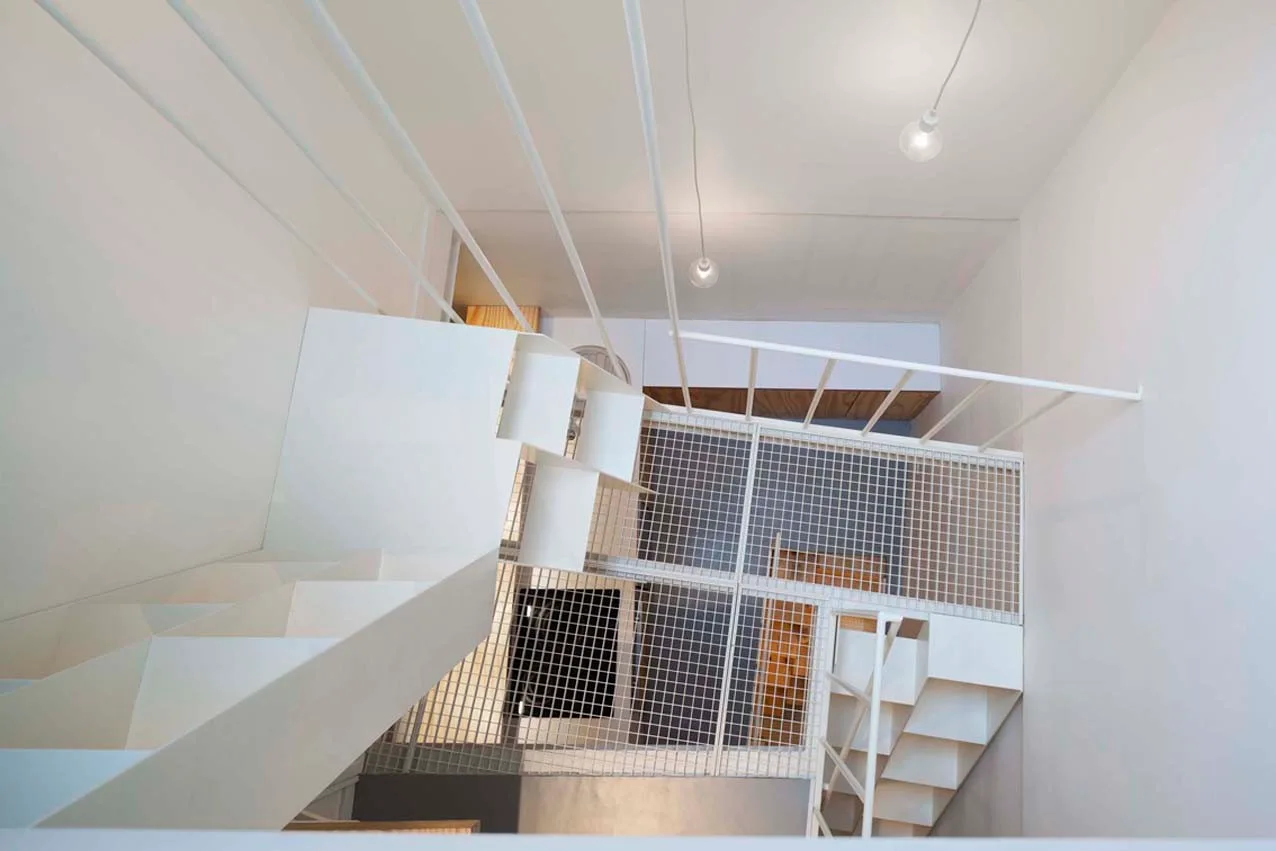 A high-angle view looking down a minimalist white metal staircase with open risers, vertical railings, and a steel mesh floor platform.