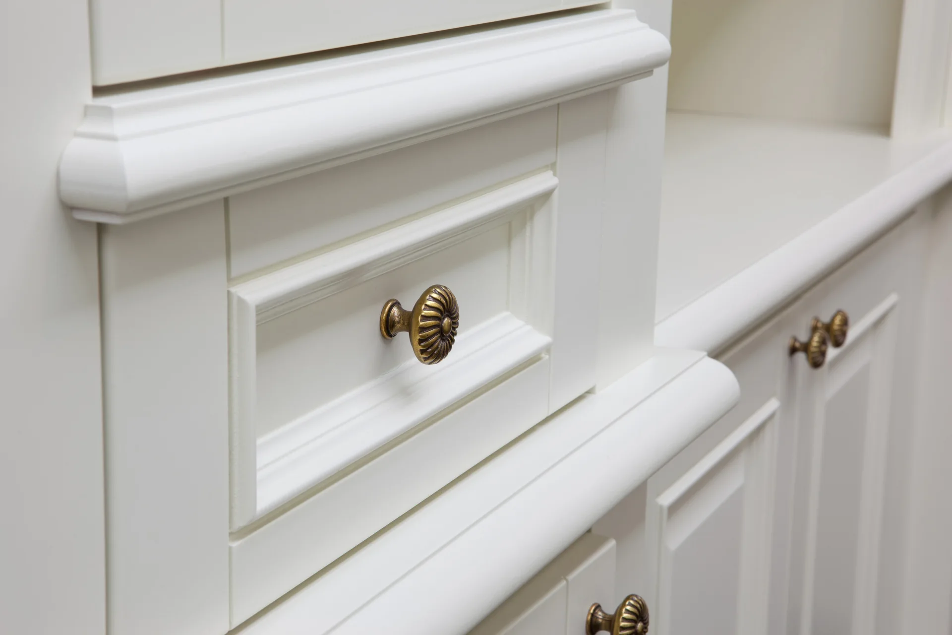 Close-up of white painted cabinetry featuring recessed drawer panels and ornate, circular antique brass knobs.