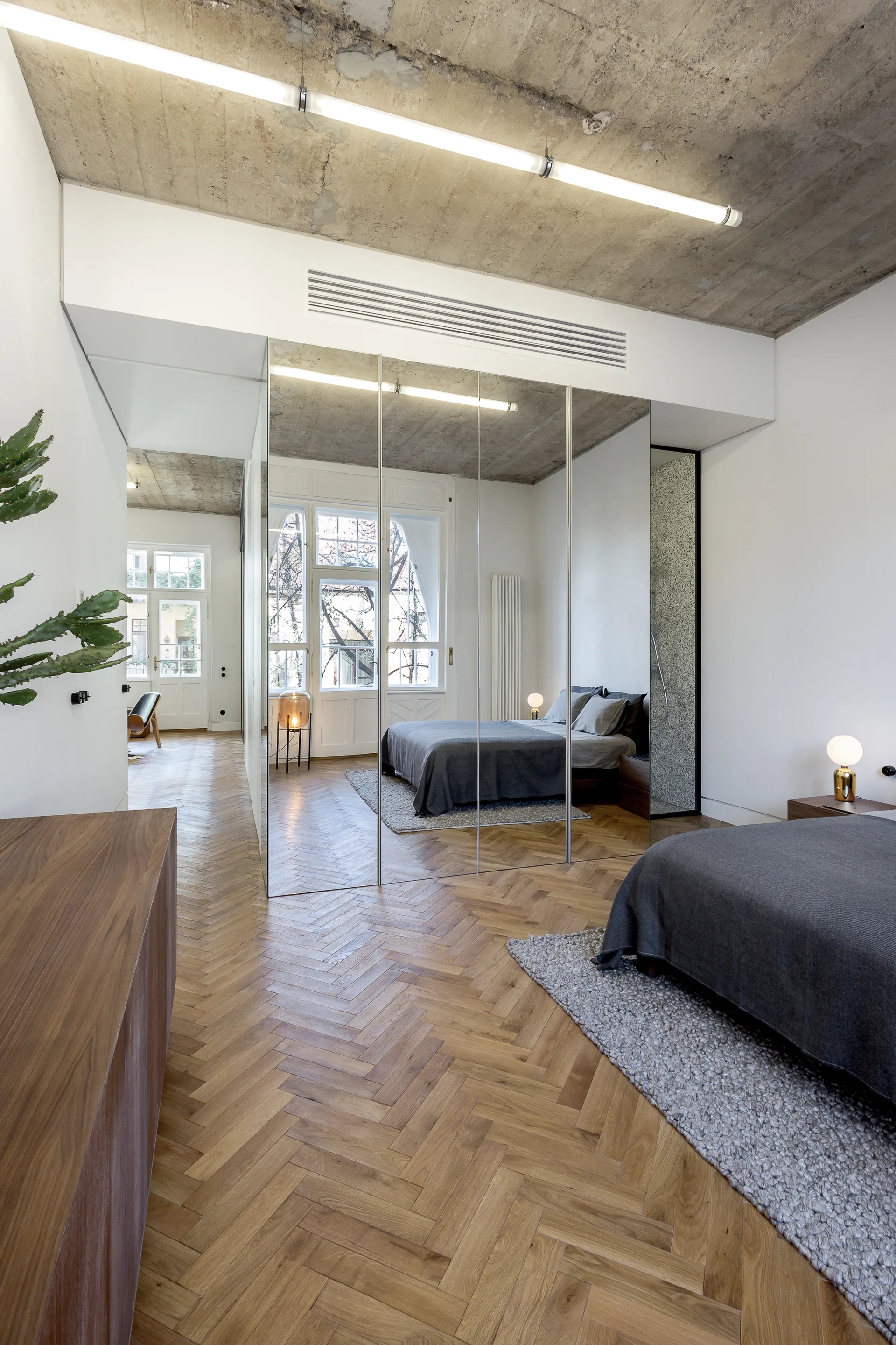 A minimalist bedroom featuring herringbone wood flooring, an exposed concrete ceiling, and floor-to-ceiling mirrored closet doors.