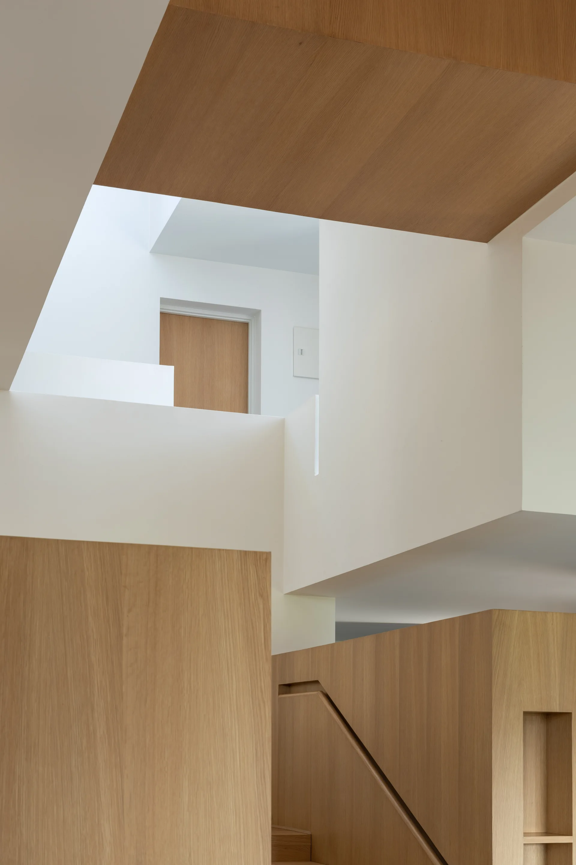 A multi-level minimalist stairwell features sharp geometric white walls contrasted with light oak wood paneling under a bright skylight.