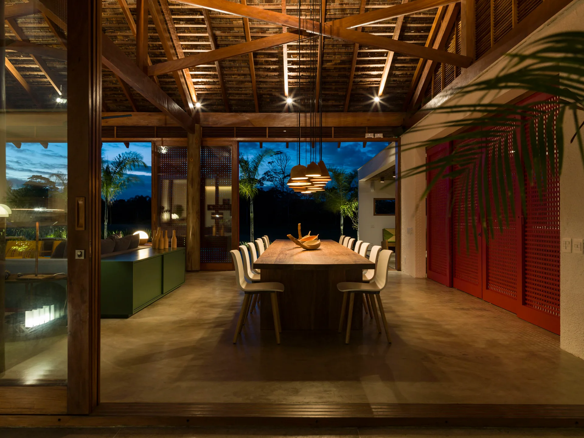 An open-air dining room features a long wooden table beneath an exposed timber ceiling, illuminated by a cluster of pendant lights at dusk.