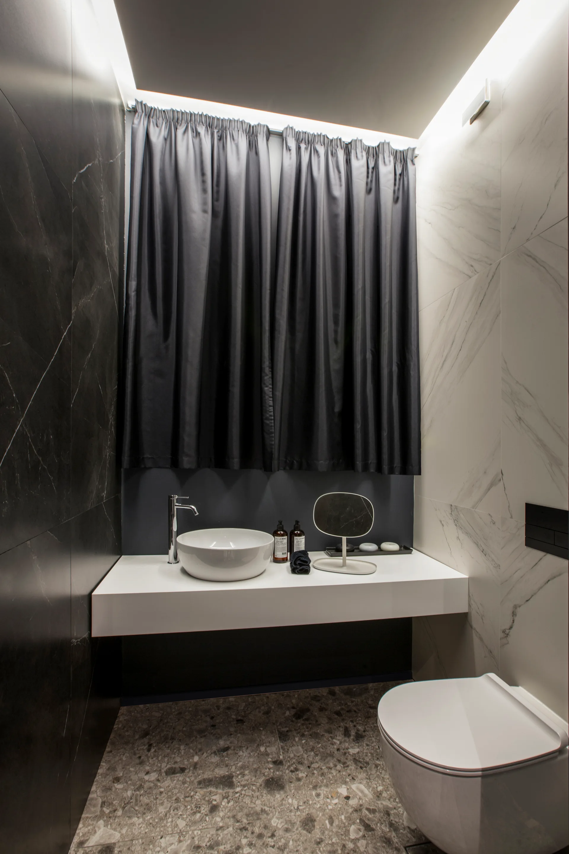 A modern bathroom features black and white marble-tiled walls, a floating white vanity with a vessel sink, and a dark grey curtain under cove lighting.