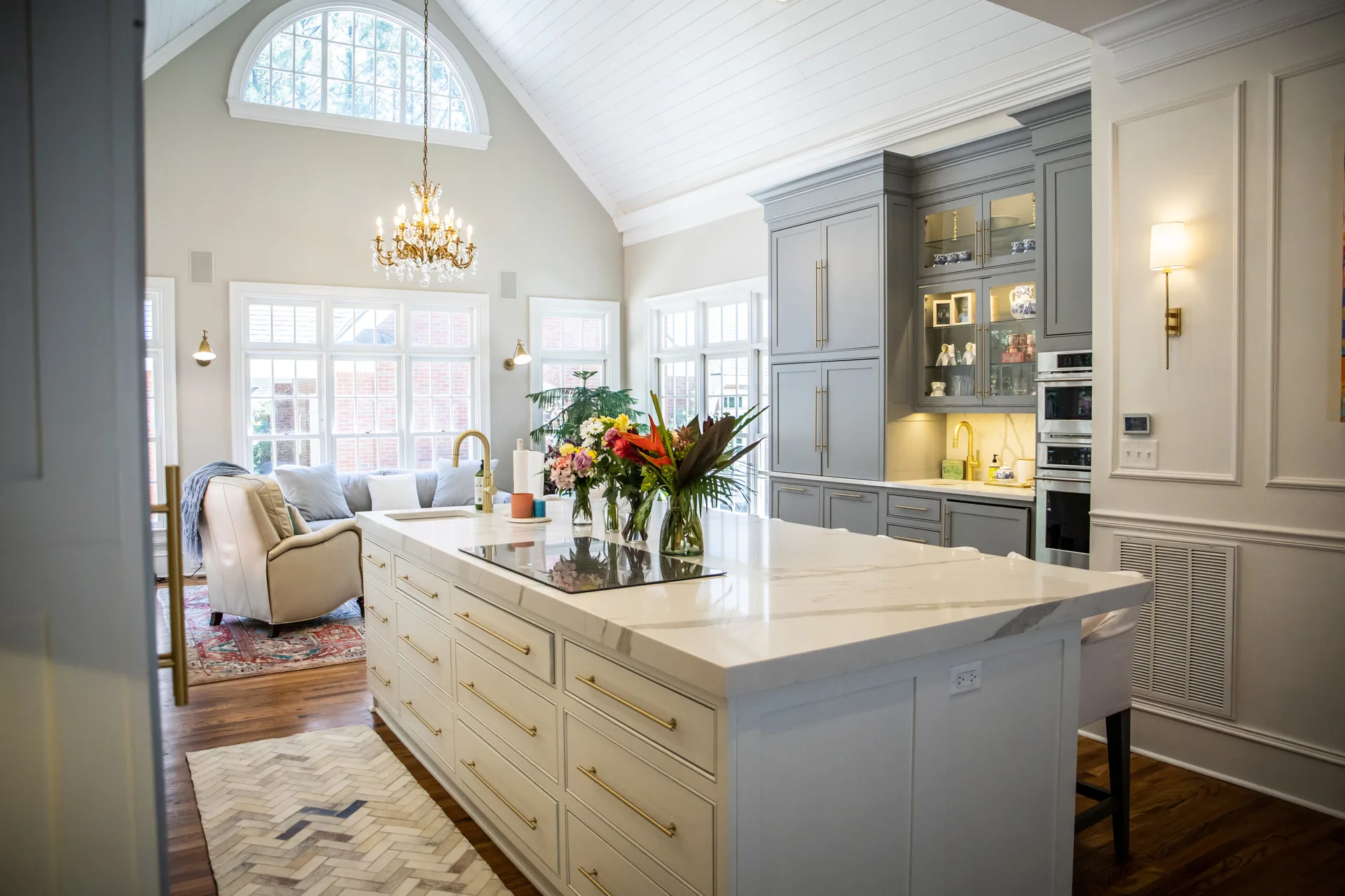 A bright transitional kitchen featuring a large white marble island with gold hardware, grey cabinets, and a high vaulted wood plank ceiling.