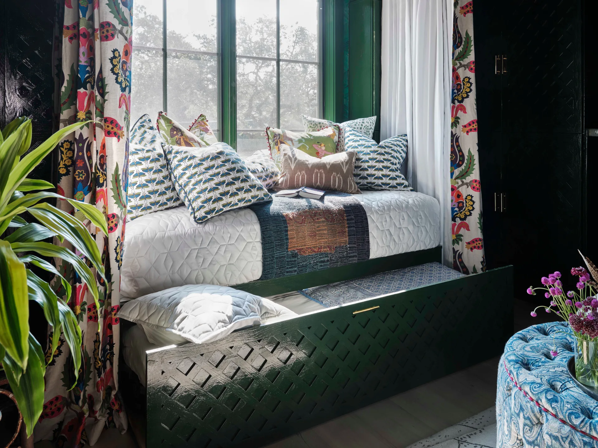 An eclectic daybed with a green lattice trundle sits under a window framed by floral curtains and colorful patterned pillows.